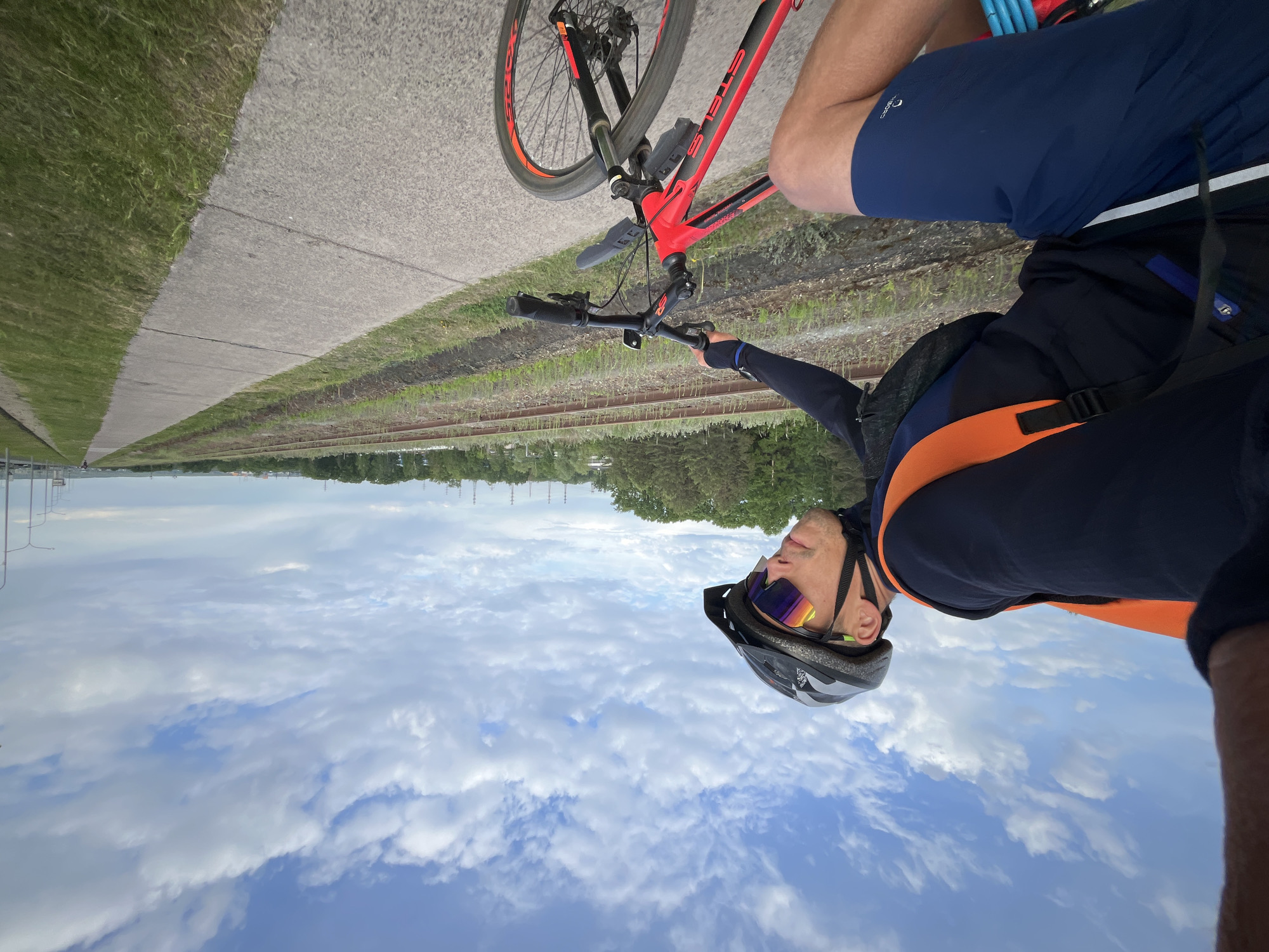 The image shows a man riding a red and black mountain bike on a paved path adjacent to railway tracks and a grassy area, likely in Chaikovsky, Russia. He is wearing a black helmet, sunglasses, and a dark blue and orange top. The sky is visible in the background with scattered clouds. The angle of the photo appears to be taken from a low angle.