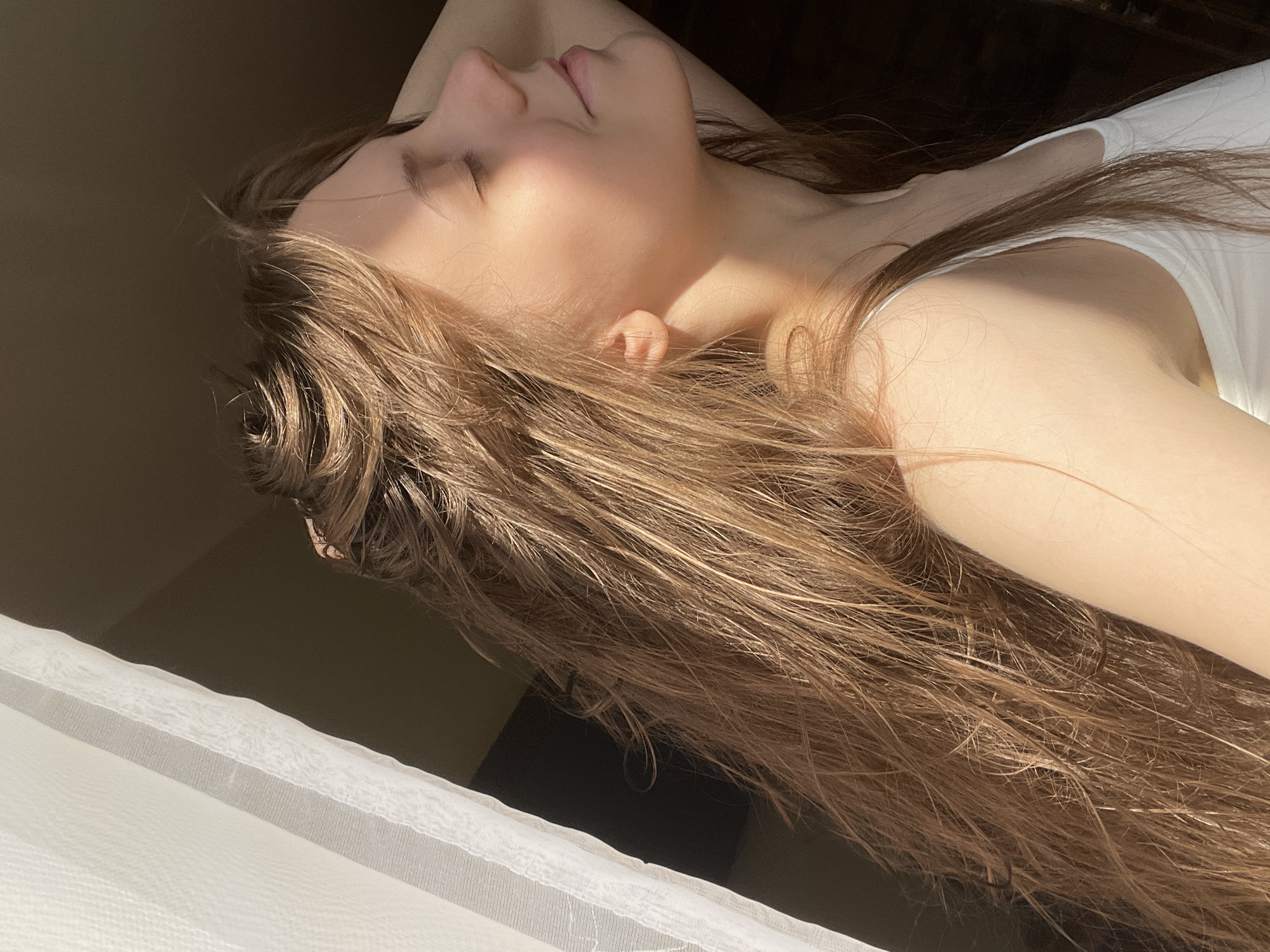 A side-profile close-up of a young woman's face and hair is captured, with the image rotated 90 degrees counter-clockwise. She is lying down, her eyes closed, with her head tilted back. Sunlight streams across her face, highlighting her nose, cheek, and forehead, creating a warm glow. Her long, light brown hair is spread out around her, some strands catching the light. She is wearing a white top, the strap of which is visible on her shoulder. The background is dark and indistinct, but a sheer white curtain is visible in the bottom left corner, suggesting an indoor setting near a window. The lighting suggests it is daytime, likely morning or late afternoon, given the direction and quality of the sunlight. The overall mood is one of peacefulness and relaxation. No text is visible in the image. The location context is Ufa, Russia.