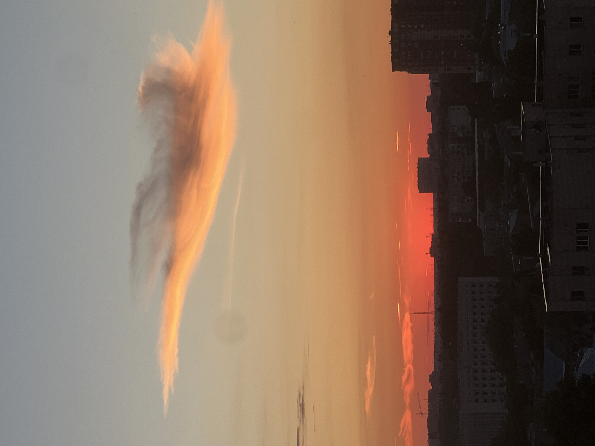 This is a photograph of a city skyline at sunset, likely taken from a high vantage point in Odesa, Ukraine. The sky is a gradient of colors, transitioning from a pale blue at the top to a vibrant orange and deep red near the horizon. A distinctive, elongated cloud formation, illuminated by the setting sun, stretches across the upper portion of the sky, appearing in shades of orange and pink.
Below the sky, the silhouette of a city is visible. In the middle ground, there are several large, multi-story buildings, some appearing residential with numerous windows, and others are more industrial or commercial in appearance. Cranes, indicative of construction, are visible in the distance on the left. In the foreground, the tops of more buildings are seen, their details obscured by the low light. The overall impression is one of an urban environment at the end of the day, with the dramatic sky providing a striking backdrop. There are no people or animals clearly discernible in the image. The atmosphere is tranquil, with the focus primarily on the natural light display and the urban landscape.