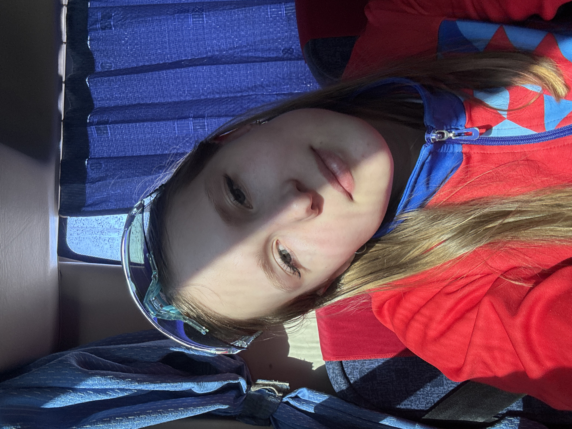 A girl with blonde hair is reclining. She is wearing a red zip-up jacket with blue accents and blue sunglasses. She is likely seated on a bus due to the blue, patterned fabric around her and the blue window blinds. Her location in Sibir', Russia, cannot be ascertained from the visual information alone.