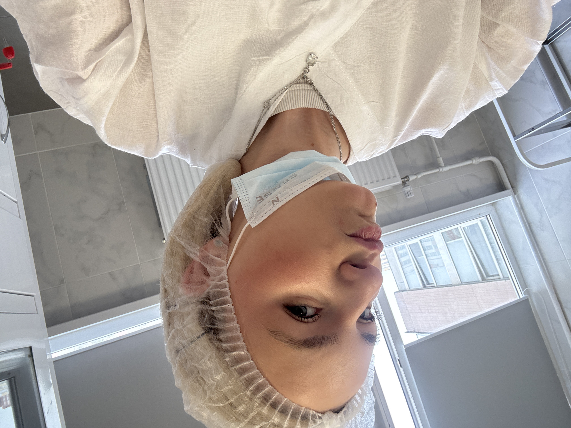 A person with fair skin, blond hair covered in a white hair net, and wearing a light blue surgical mask is shown from a low angle, looking down at the camera. They are wearing a white collared shirt with a silver necklace. The background includes a white tiled wall, a white radiator, a window looking out onto a cityscape featuring buildings in Saint Petersburg, Russia. Also, in the background is a white clothing rack.