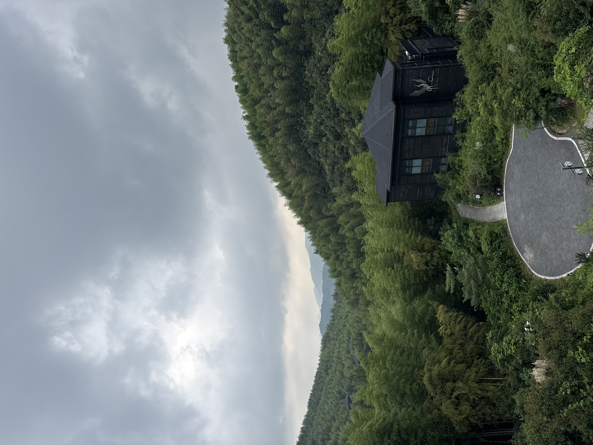 A dark, modern house with a pitched roof sits nestled among lush green bamboo forests on a hillside. The house features dark wood siding and large windows. A pathway leads to the front of the house, which is surrounded by dense vegetation. In the background, rolling hills covered in more bamboo extend into the distance under a cloudy sky. The scene appears to be a tranquil, secluded natural setting, possibly a resort or private residence, located in the Wuxing District of China.