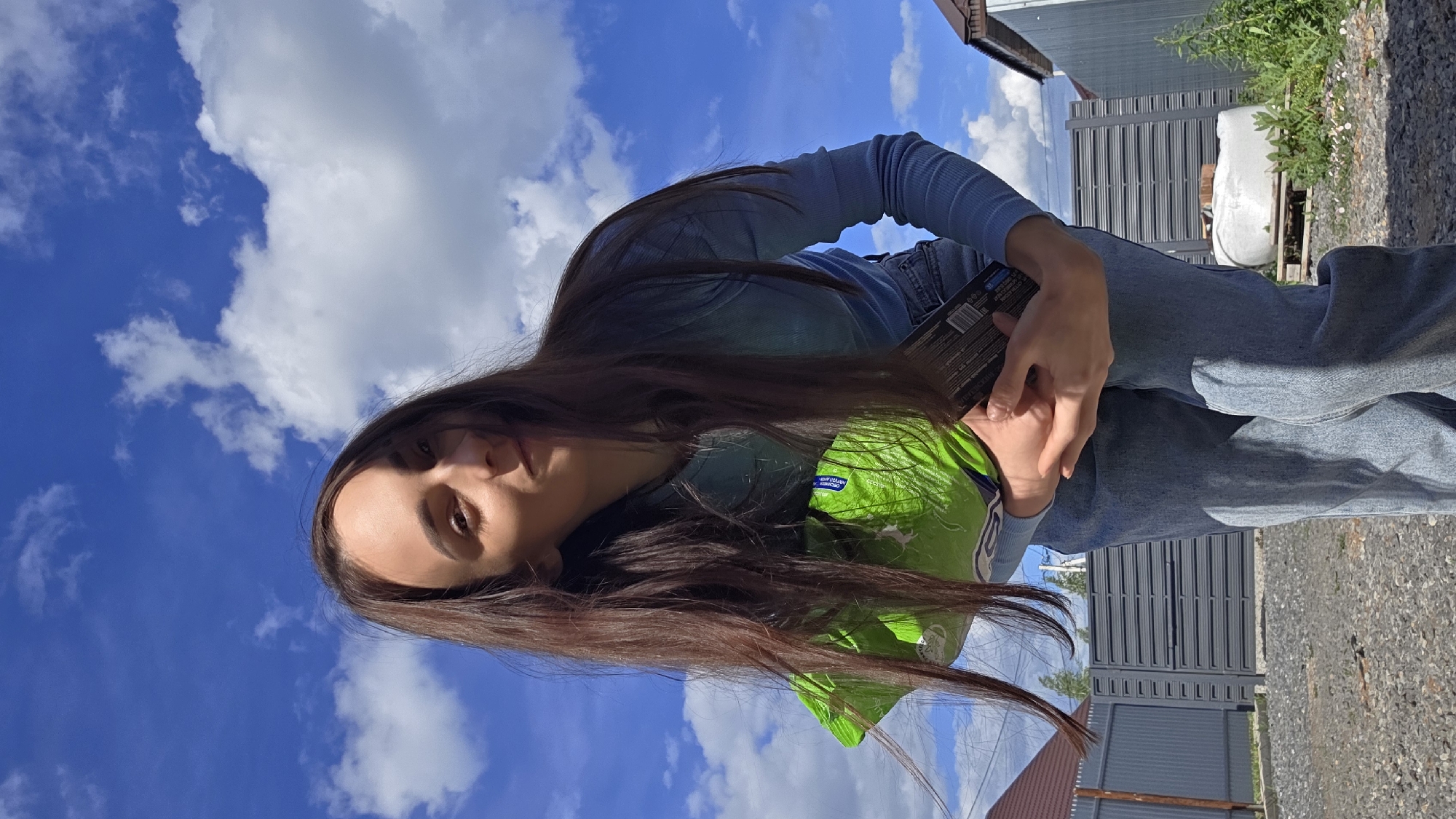 A young woman with long, dark hair is holding a bag of Lay's potato chips and a dark object (possibly a book or another snack) while standing outside. She is wearing a blue long-sleeved shirt and jeans. The background shows a bright blue sky with scattered white clouds, a building with gray metal siding, some greenery, and a gravel ground. The image appears to be taken outdoors in daylight. Bulgakovo is not evident in the image.