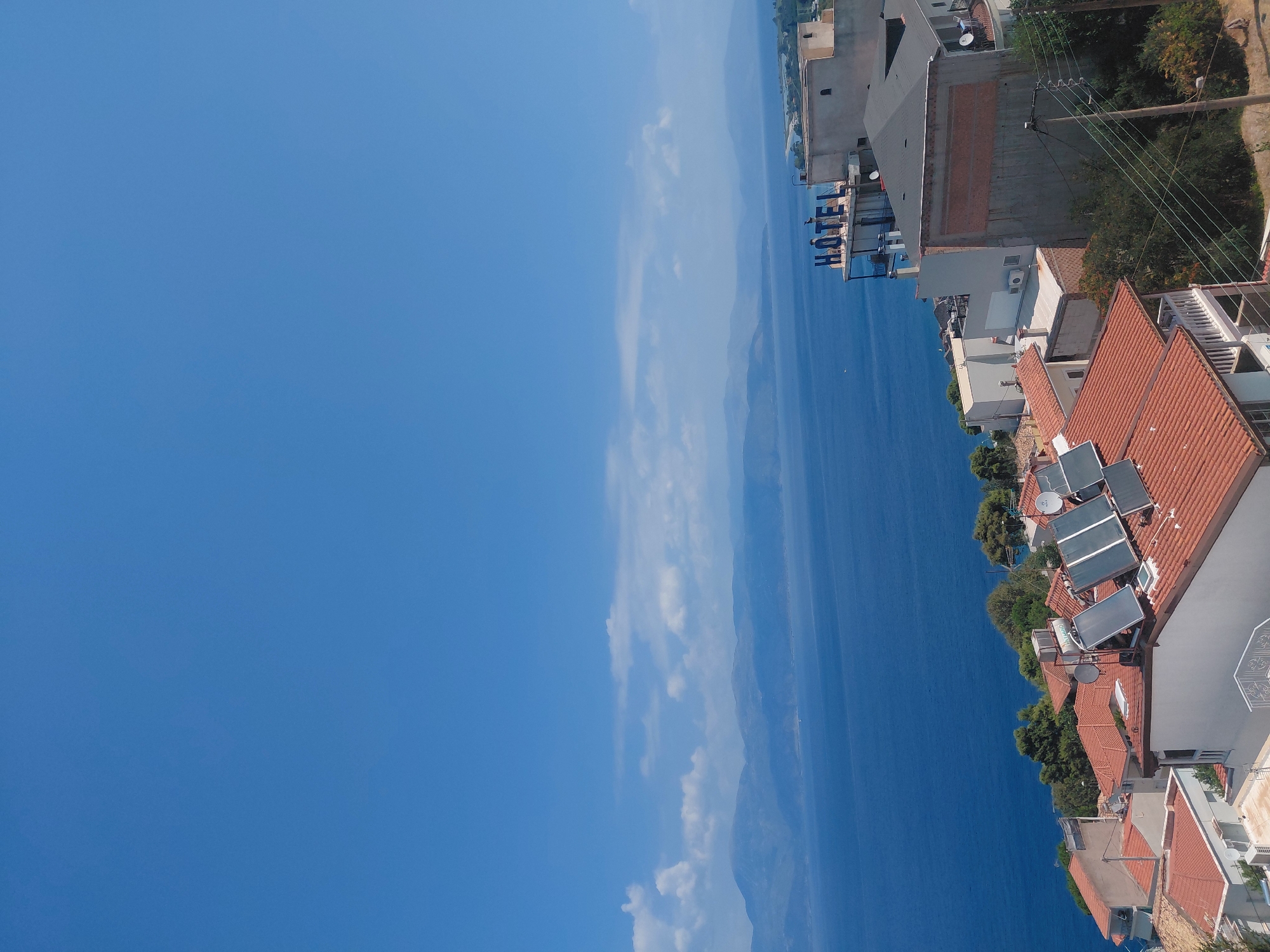 This is a high-angle, full shot image of a coastal town under a clear blue sky. The foreground is dominated by the terracotta tiled roofs of several houses, some equipped with solar panels and air conditioning units. A few trees are interspersed among the buildings. In the midground, a larger building with a prominent "HOTEL" sign is visible, situated on a slope overlooking the sea. The sea itself is a deep blue, extending towards a hazy horizon where distant mountains are silhouetted against the sky. The location is the Municipal Unit of Aegio, Greece. There are no people visible in the image.