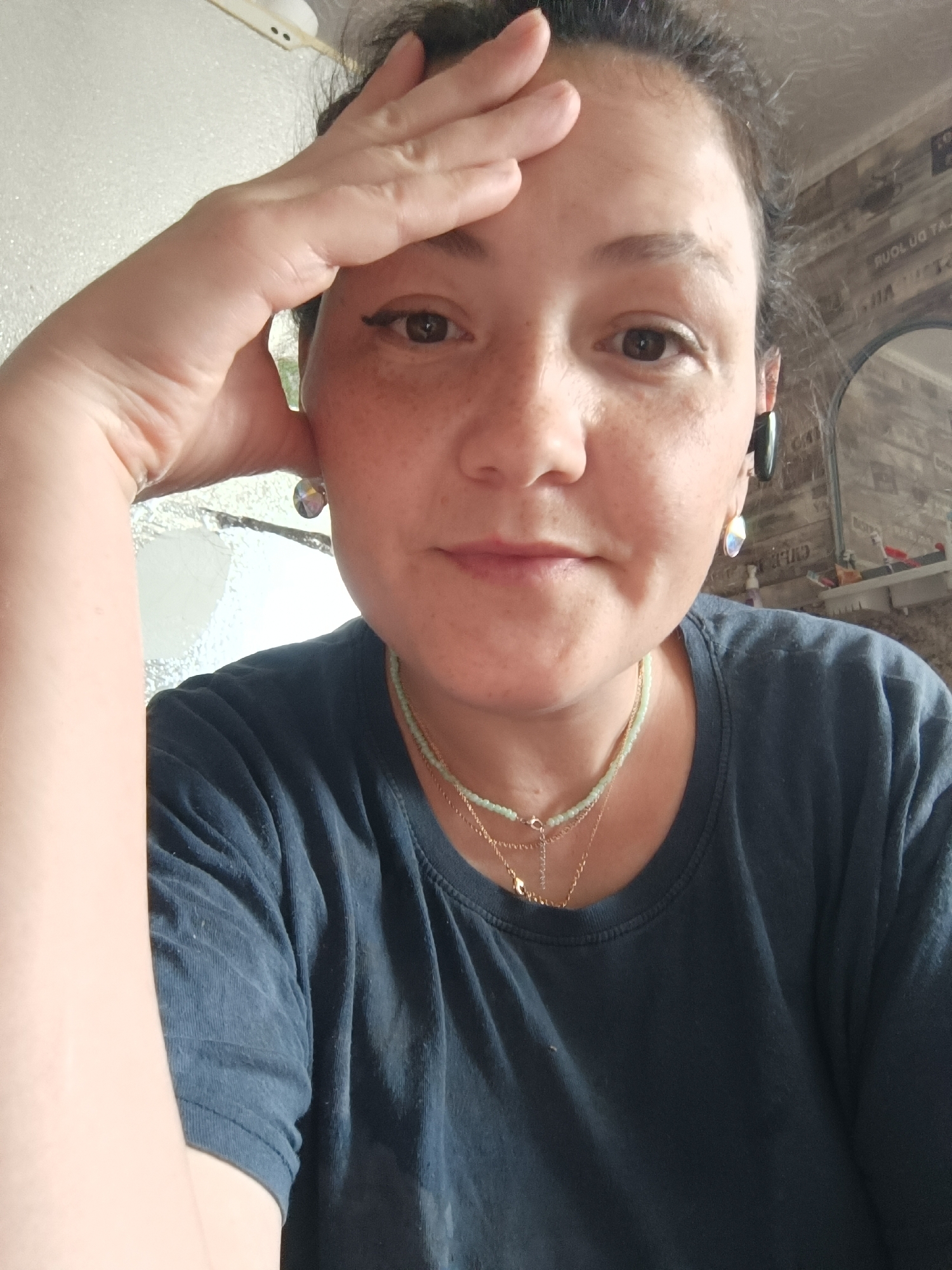 A woman is captured in a close-up, selfie-style shot, holding her right hand to her forehead. She is wearing a dark blue t-shirt and multiple necklaces, including one with small green beads and a delicate gold chain with a pendant. She also has a dark earbud in her right ear and a large, multi-faceted earring in her left ear. Her hair is pulled back, and her face is turned slightly towards the camera. Freckles are visible across her nose and cheeks. The background is blurred, but suggests an indoor environment with a wall featuring a wood-grain pattern. The lighting is soft, illuminating her face and upper body. There is no information in the image to suggest it is located in Karsha, Russia, or any other specific location.