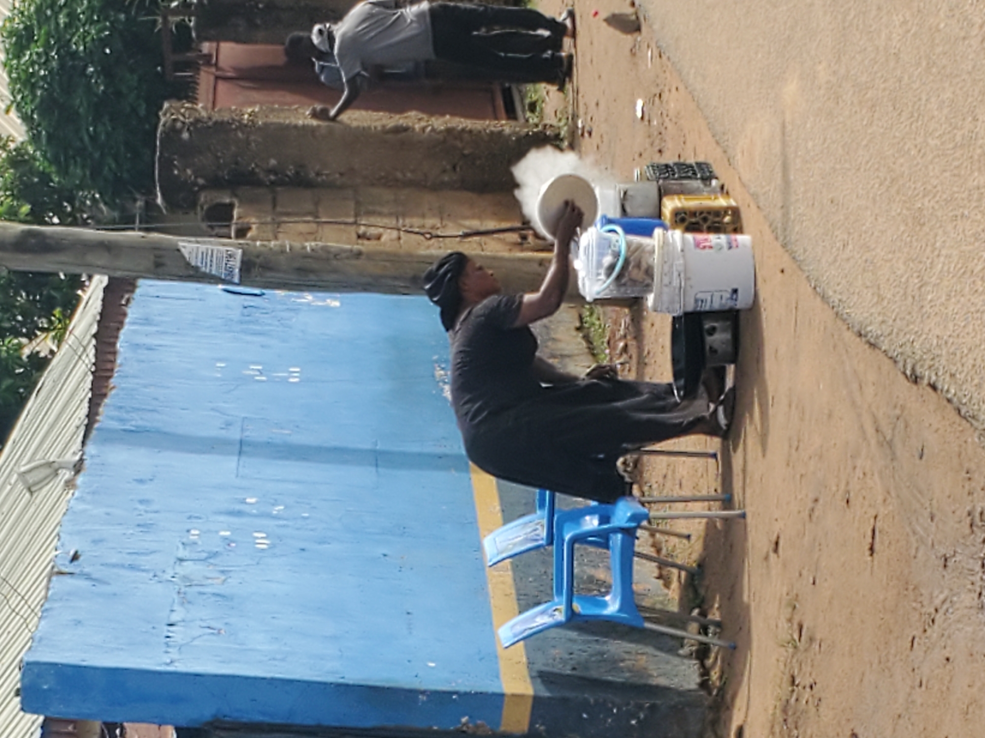 In a dusty outdoor area, a woman dressed in a black headscarf and dress is seen tending to cooking pots on the ground. She is holding a white plate and appears to be cooking, as evidenced by the steam rising from a pot. In front of her are several containers, including a white bucket filled with items that resemble fish and another blue bucket. Two blue plastic chairs are positioned against a vibrant blue wall, which has a yellow stripe near its base. To the right, two men are standing near a brick structure and a reddish-brown metal gate. One man is wearing a grey shirt and dark pants, while the other is in a grey t-shirt and black pants, with a white cap. The environment suggests a roadside scene, possibly in the city of Jos, Nigeria, characterized by the unpaved ground, the building materials, and the general atmosphere.