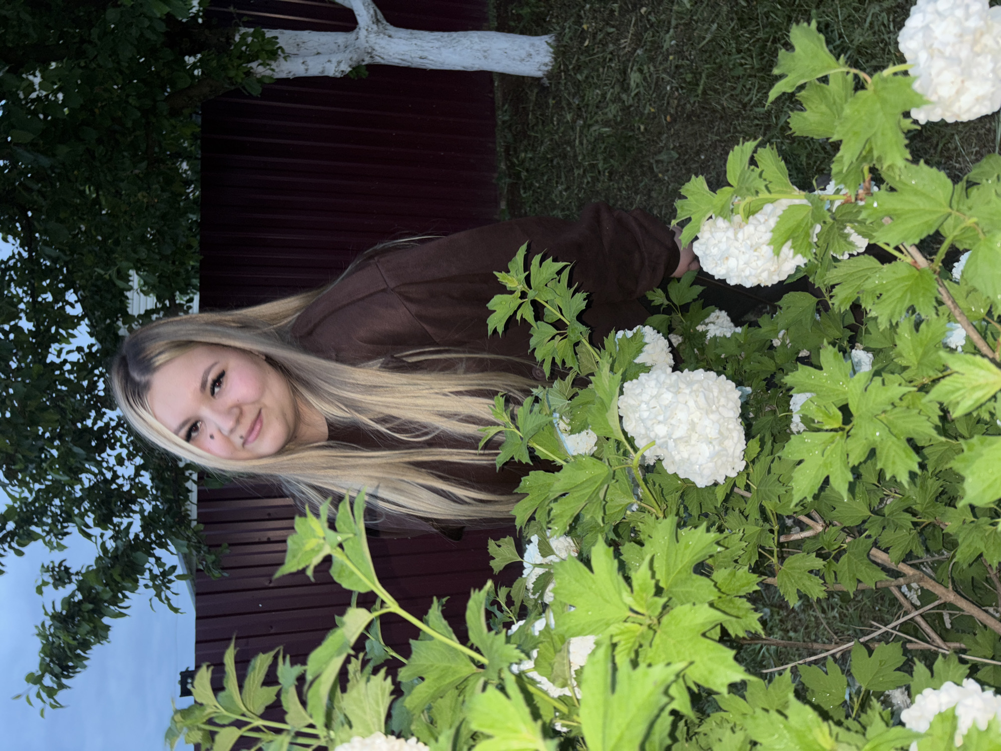 A blonde woman in a brown hooded shirt smiles at the camera, half obscured by a bush with large white snowball-shaped flowers. The bush is primarily green, with water droplets on the leaves. Behind the woman is a red fence and a white painted tree trunk. The photo appears to be taken outdoors, at dusk or night. The location is in Фоки, Russia.
