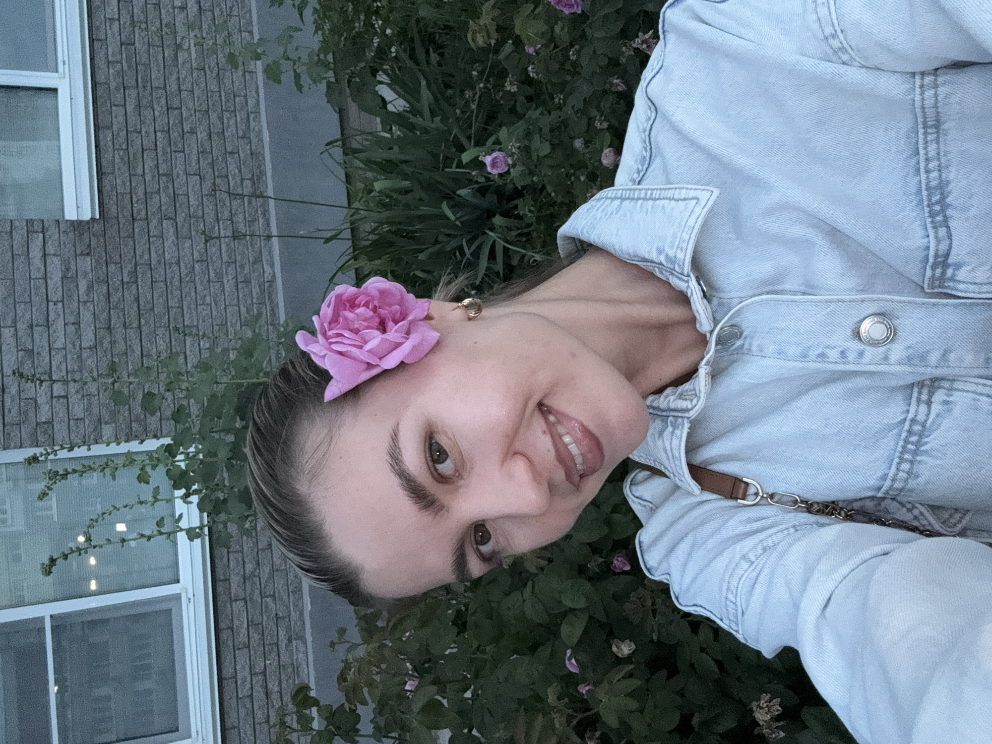 A medium shot shows a young woman with a pink rose tucked behind her right ear. She is smiling and looking at the camera. She is wearing a light blue denim shirt and a brown strap from a bag is visible on her shoulder. In the background, there is a brick building with a window and a dense green bush with some small pink flowers. The overall lighting suggests it is either early morning or late evening. The location appears to be outdoors, likely a residential area.
