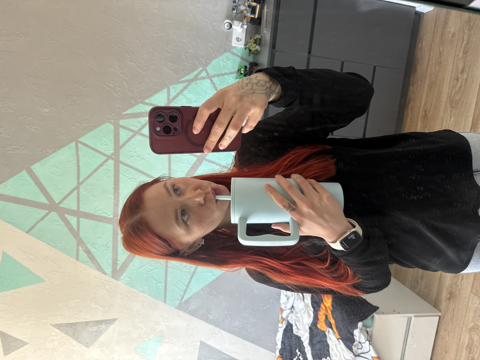 A person with red hair is reflected in a mirror, taking a selfie while drinking from a light blue cup with a straw. The person is wearing a black top and has a smartwatch on their left wrist. They are holding a maroon smartphone in their right hand. The reflection also shows part of a gray and white patterned wall and a gray cabinet or dresser. Several Lego or similar toy vehicles are visible on the cabinet. The environment includes a wooden floor and a patterned blanket or sheet near the bottom of the frame. There is no information provided in the image that could be used to confirm the location is in Rostov-on-Don, Russia.