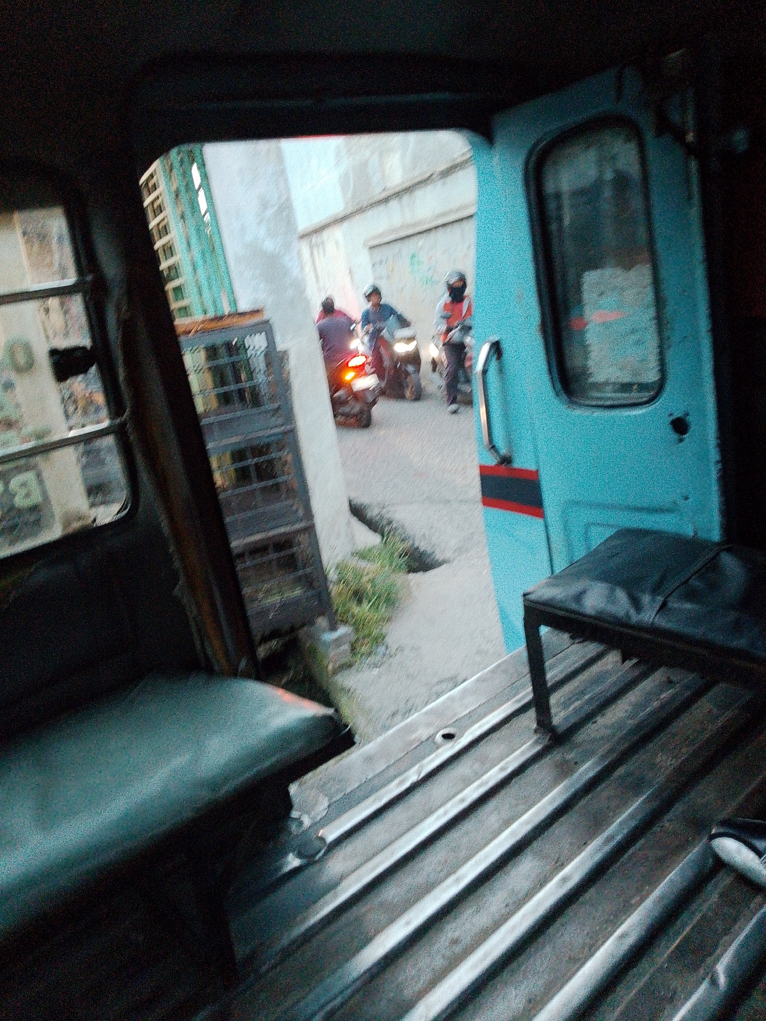 The image captures a view from inside a vehicle, looking out through an open doorway. The interior of the vehicle is visible, featuring dark-colored seats and a metallic floor with parallel ridges. Outside, a street scene unfolds. Two individuals on motorcycles are present. One motorcycle has its brake light illuminated. The road appears to be paved, with some patches of vegetation along the side. A building with a light-colored facade stands in the background, with a green structure adjacent to it. A teal-colored door with a metal handle and a red stripe is visible on the right side of the frame, forming part of the vehicle's exterior. The scene suggests a moment captured in an urban environment, possibly Bandar Khalipah, Indonesia, given the context.