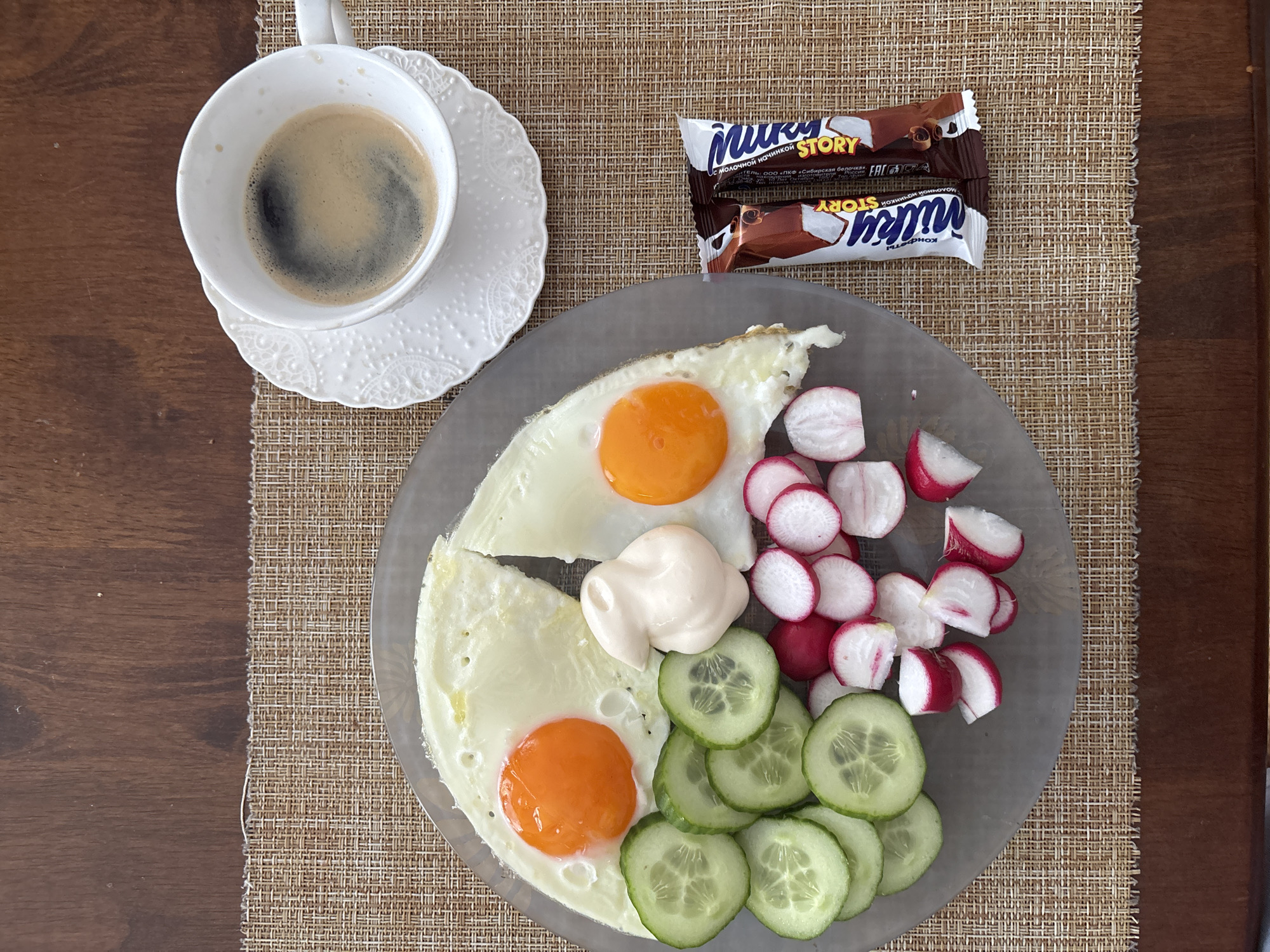 A top-down view shows a breakfast spread on a textured placemat over a dark wooden table. The meal consists of a plate with two fried eggs, sliced cucumbers, sliced radishes, and a dollop of mayonnaise. To the right of the plate is a white teacup filled with dark coffee, resting on a decorative white saucer. Two chocolate candy bars, identified as "Milky Story," are placed next to the teacup. The scene does not provide any information about the city of Sochi, Russia, or any people present.