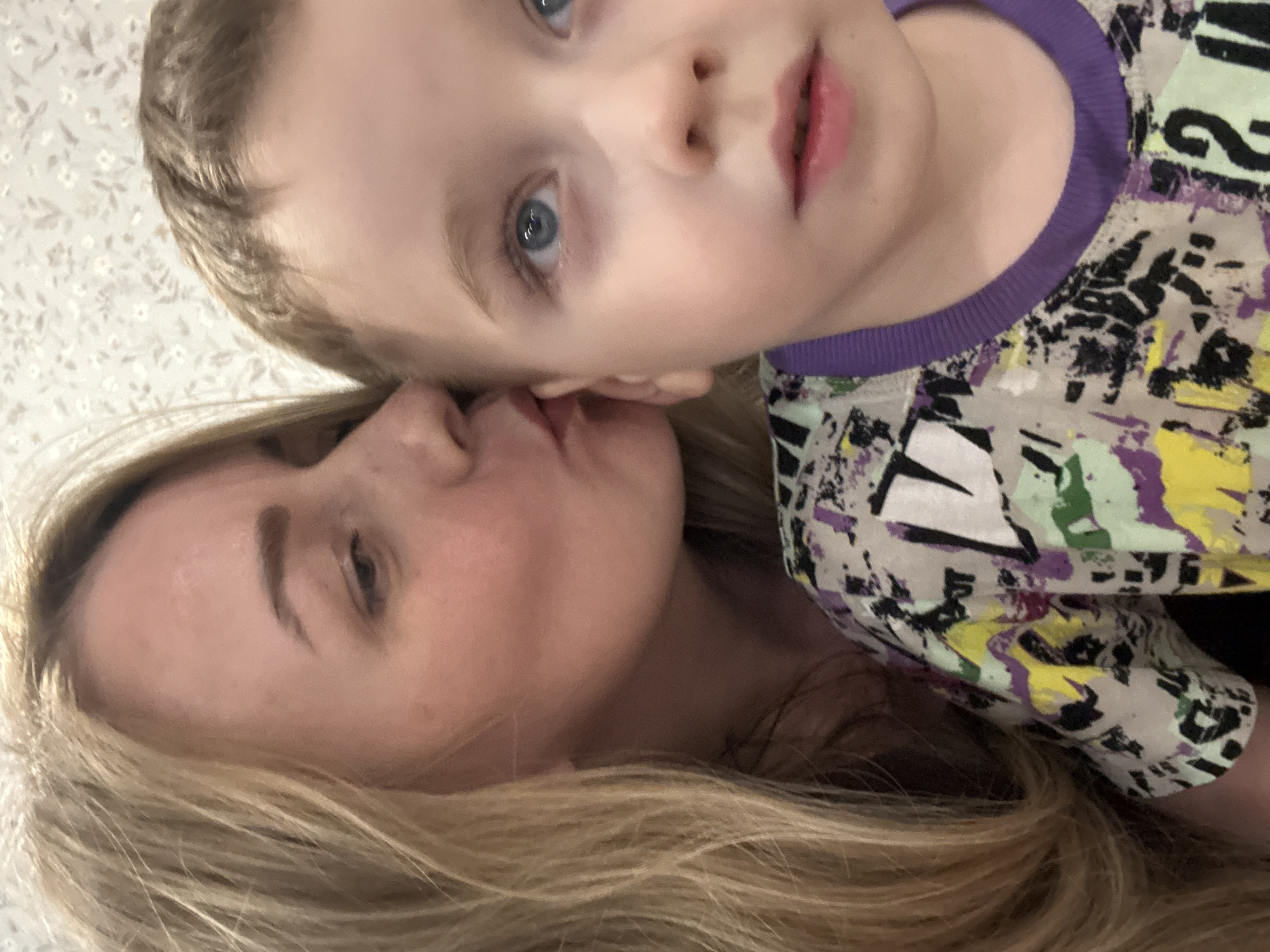 A fair-skinned woman with blonde hair and closed eyes kisses the cheek of a fair-skinned young child with blue eyes, a fair complexion, and blonde hair. The child is wearing a gray, black, purple, and yellow patterned shirt with a purple collar. The background appears to be a light-colored wall with a pattern. The location is not discernible from the image.