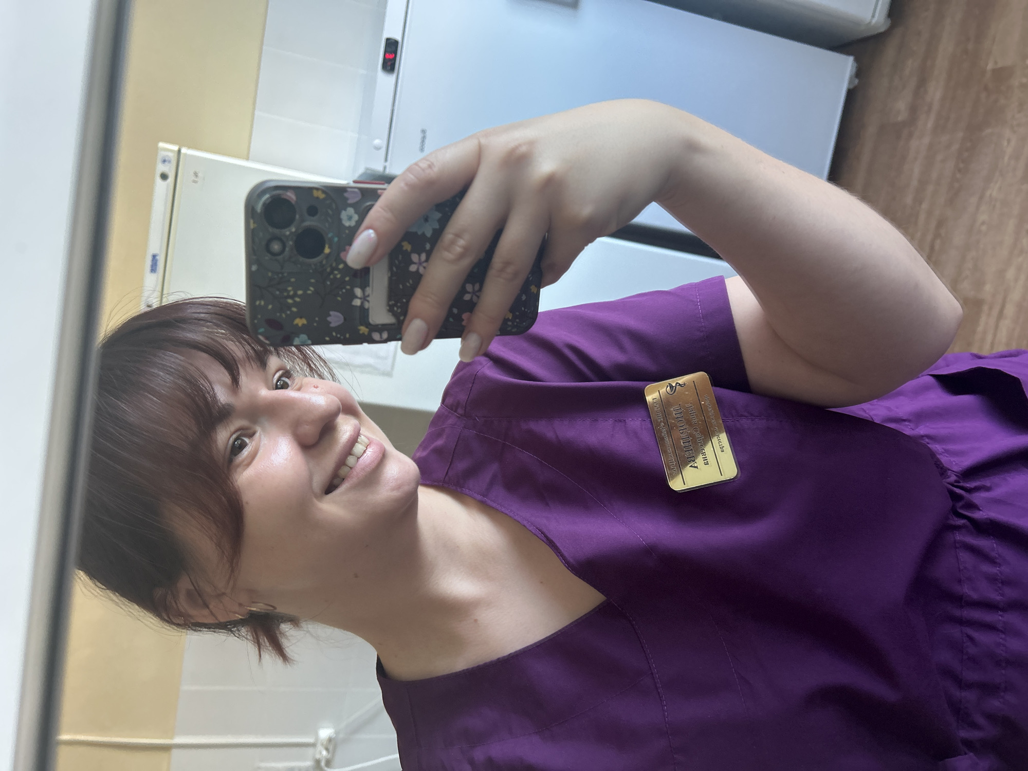A young woman with brown hair and bangs, wearing a purple scrub top and a golden name tag, takes a mirror selfie. She is smiling, holding a floral-patterned smartphone in her right hand. The background appears to be an indoor setting with white tiled walls and what seem to be refrigerators. The image is captured in a reflection, indicating the presence of a mirror. The name tag on her uniform suggests she is likely in a medical or healthcare setting. The ambient lighting suggests it is daytime. There are no other individuals or animals visible in the image. The overall scene is clean and appears to be a private moment captured by the subject.
