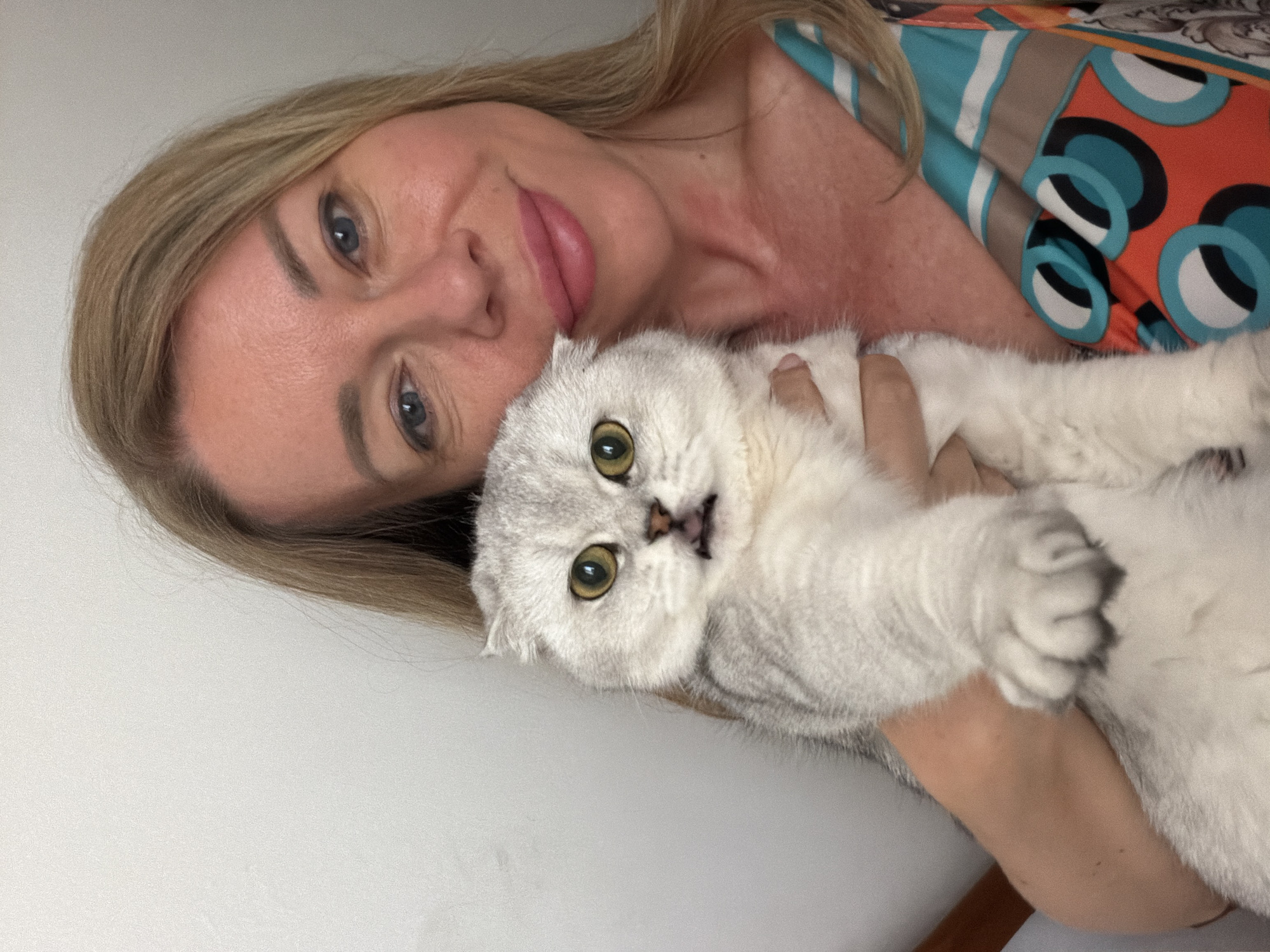 A woman with blonde hair and blue eyes is holding a silver Scottish Fold cat. The cat has green eyes and its mouth is slightly open, revealing its tongue. The woman is wearing a colorful dress with a patterned neckline. The background is a plain white wall. The image is a selfie, with the woman holding the camera at an arm's length. There is no indication of the location being Rome, Italy.