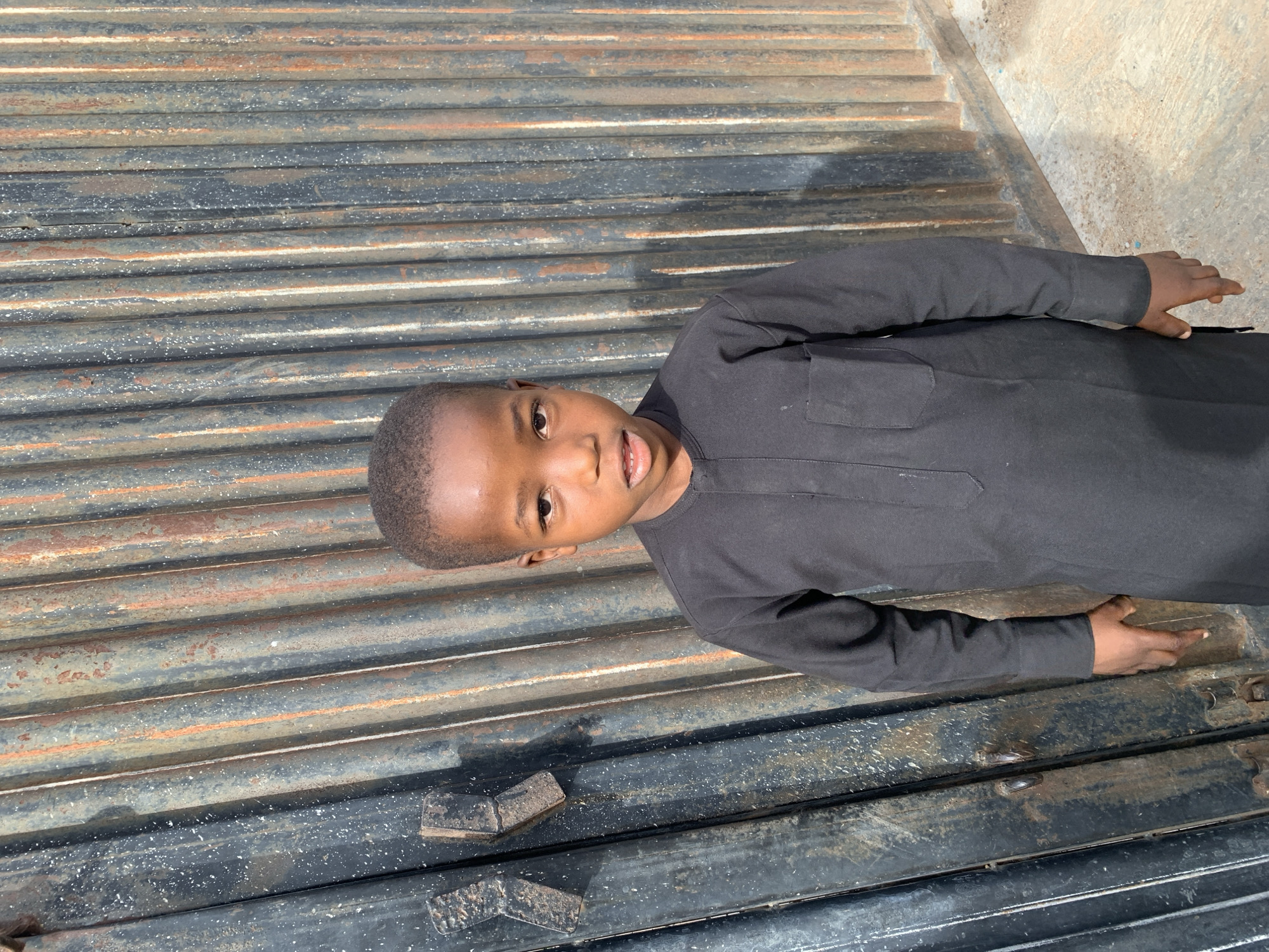 A young boy, dressed in a dark, long-sleeved shirt, is lying on a corrugated metal surface. The metal has a combination of dark and rusty hues. The boy is looking directly at the camera with a slight smile. The location is not discernible from the image. The city of Jos, Nigeria, is not discernible from the image.