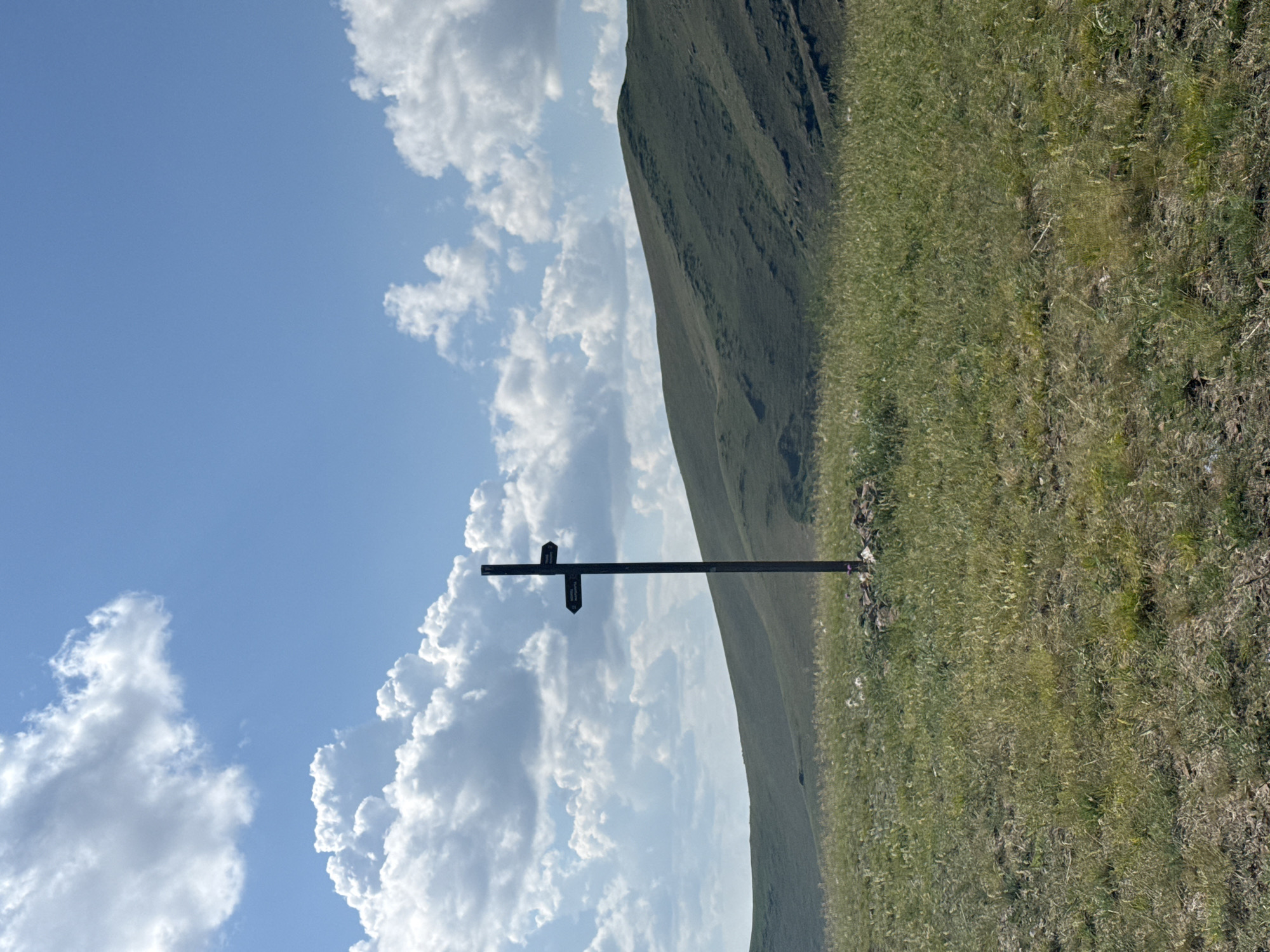 A dark, cross-shaped signpost stands in the center of a grassy, rolling landscape under a partly cloudy blue sky. The signpost has two directional arrows, one pointing left and the other pointing right. The landscape consists of green hills in the background and a field of tall, dry grass in the foreground. A small pile of rocks surrounds the base of the signpost. The scene appears to be outdoors in a rural or natural setting, possibly a hiking trail, and does not contain any people or discernible structures that would suggest it is within the city limits of Ust'-Abakan, Russia. The image is a still, clear photograph taken from ground level.