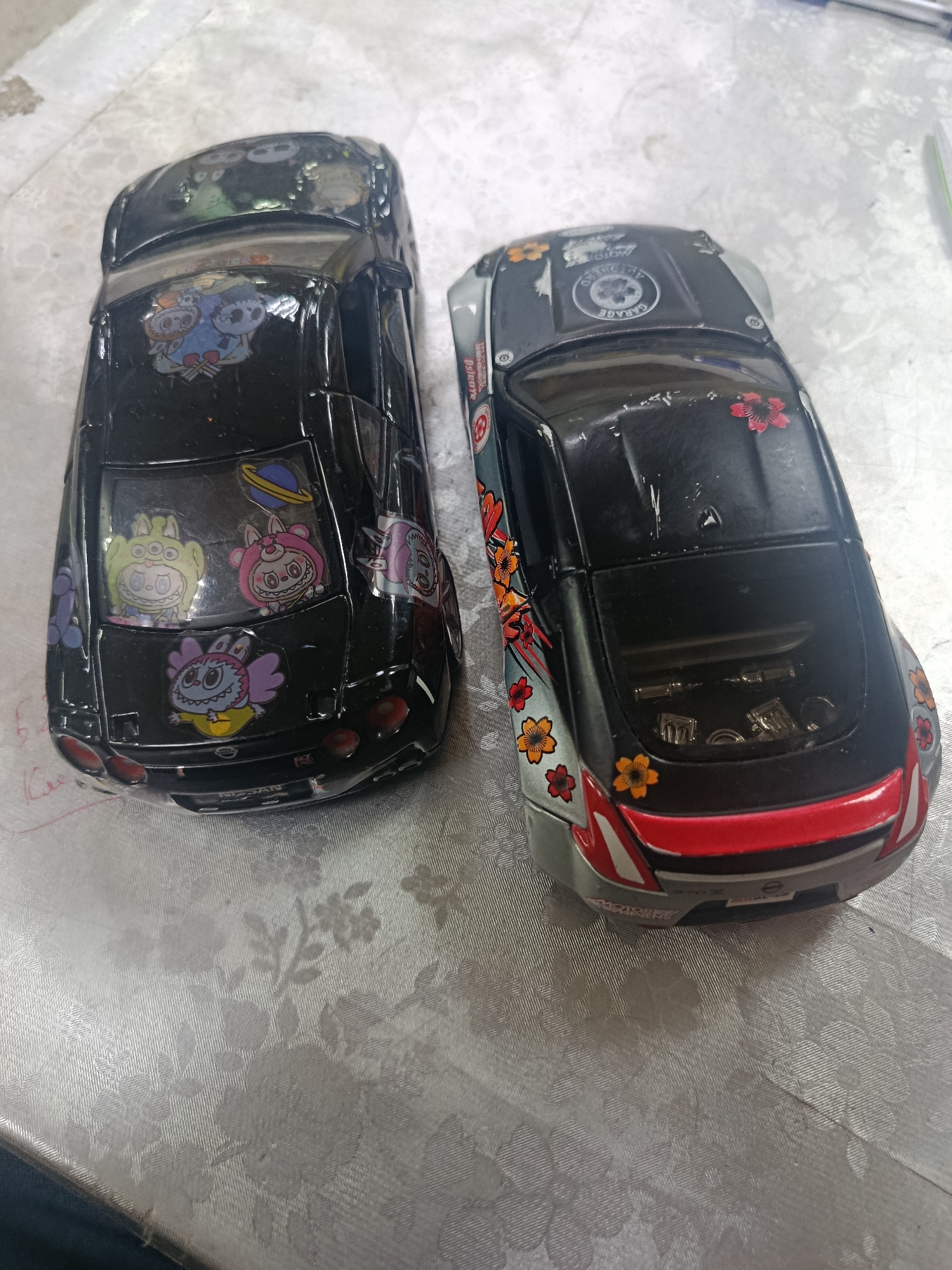 Two toy cars, a black Nissan GT-R and a silver and black Nissan 370Z, are positioned on a textured white tablecloth. Both cars are adorned with stickers. The Nissan GT-R has cartoon character stickers on its rear window and roof, including a planet. The Nissan 370Z features floral stickers and a circular logo on its roof. The tablecloth has a raised floral pattern. The image does not provide any information about the city of Sertolovo, Russia.