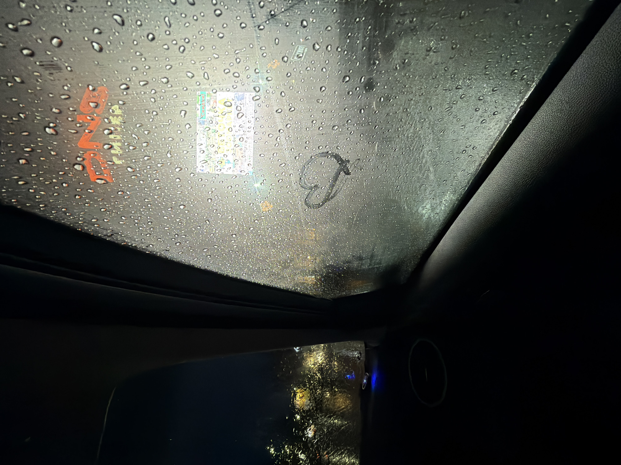 The image depicts a view from inside a vehicle through a sunroof covered in raindrops. Outside the sunroof, blurred city lights are visible, along with what appears to be a sticker or sign on the glass. A heart shape is drawn or smudged on the glass. The vehicle's interior is dark. In the bottom right corner, a circular vent and the dashboard are barely discernible. The orange reflection on the window says "ЧАЙКОВСКИЙ", which transliterates to Chaikovsky, a city in Russia. A colorful sticker with the word "СКИДКИ!" (Discounts!) is visible.