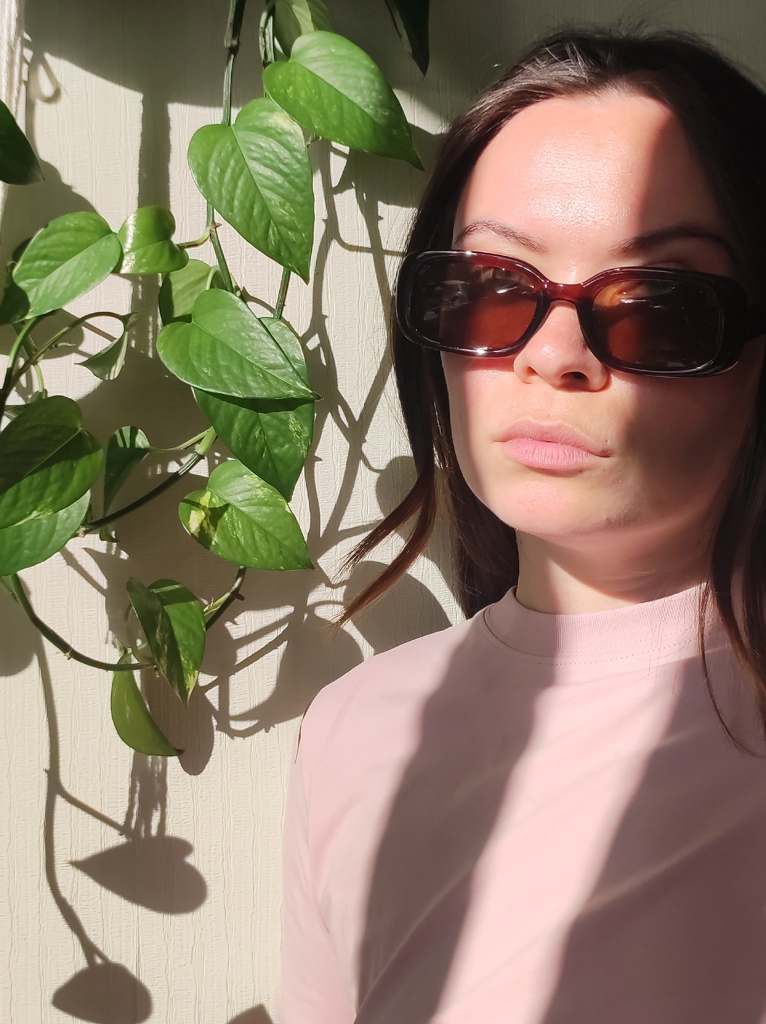 A woman with long, dark brown hair is standing near a wall indoors. She is wearing large, rectangular brown sunglasses and a light pink t-shirt. Sunlight streams in, casting shadows of the leaves from a green, leafy plant on the wall behind her. The plant appears to be a pothos. The wall is off-white with a slightly textured surface. The image does not provide enough details to confirm it was taken in the city of Chaikovsky, Russia.