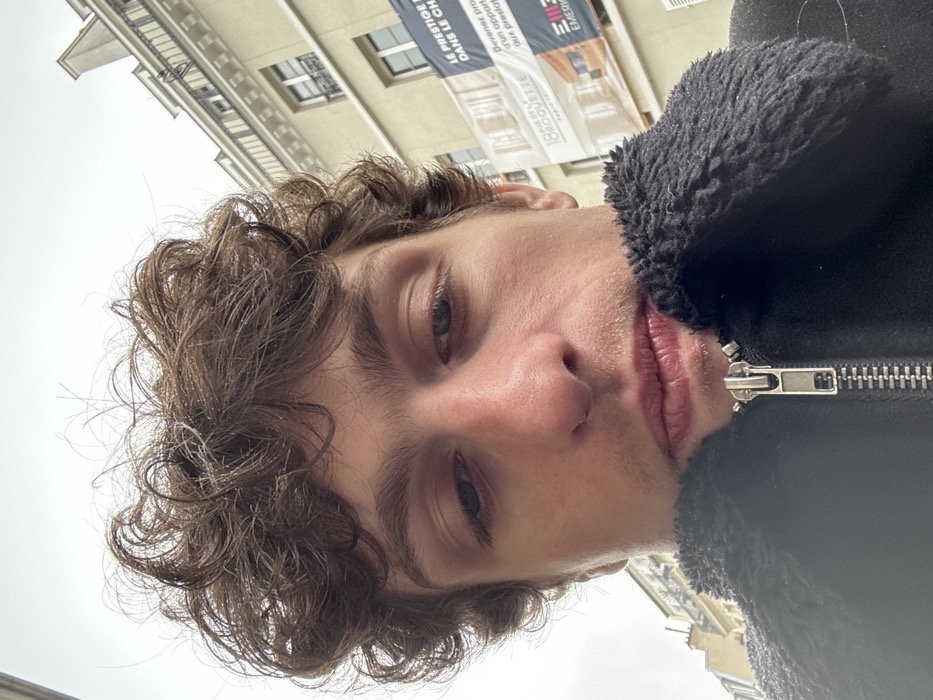The image is a low-angle, close-up selfie portrait of a male individual. He has brown, curly hair and partially visible blue eyes, gazing downwards. His face features a prominent nose, full lips, and light stubble. He is wearing a black jacket with a dark, textured collar and a visible silver zipper. The background shows an overcast sky above a light-colored, multi-story building of Haussmannian architectural style, complete with cornices and windows. A large advertising banner is affixed to the building on the right side of the frame. Text visible on the banner includes "18 RUE DE LA ROQUETTE PARIS 11e," definitively placing the scene at 18 Rue de la Roquette in the 11th arrondissement of Paris, France. Additional partial text on the banner indicates a real estate promotion, with phrases such as "LE PRESTIGE..." and "EMERIC PROMOTION."