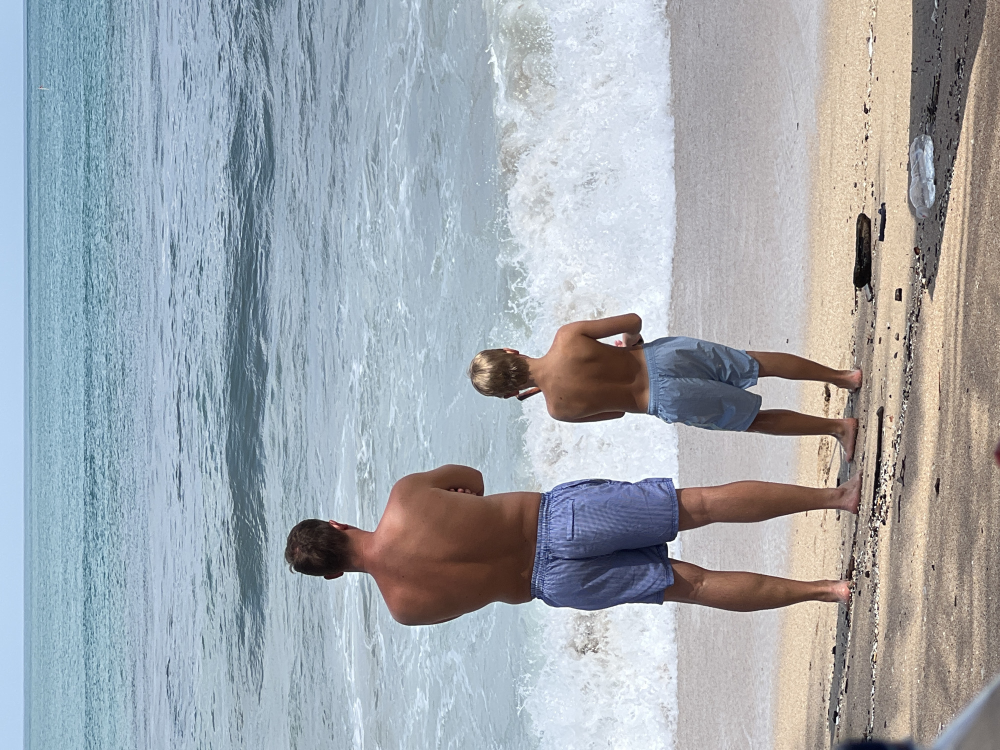 Two males, an adult and a child, stand on a sandy beach facing the ocean. The adult, on the left, is shirtless and wearing blue and white striped swim trunks. He has his arms crossed. The child, on the right, is also shirtless and wearing light blue swim trunks. He is facing away from the camera, with his left arm bent and his hand near his head. The ocean in front of them is blue with white-capped waves. The sky is overcast. The beach appears to be composed of fine sand with scattered debris, including a piece of driftwood and a clear plastic object. The scene captures a moment of observation of the sea, suggesting a relaxed or contemplative atmosphere on a beach in Kuta, Indonesia.