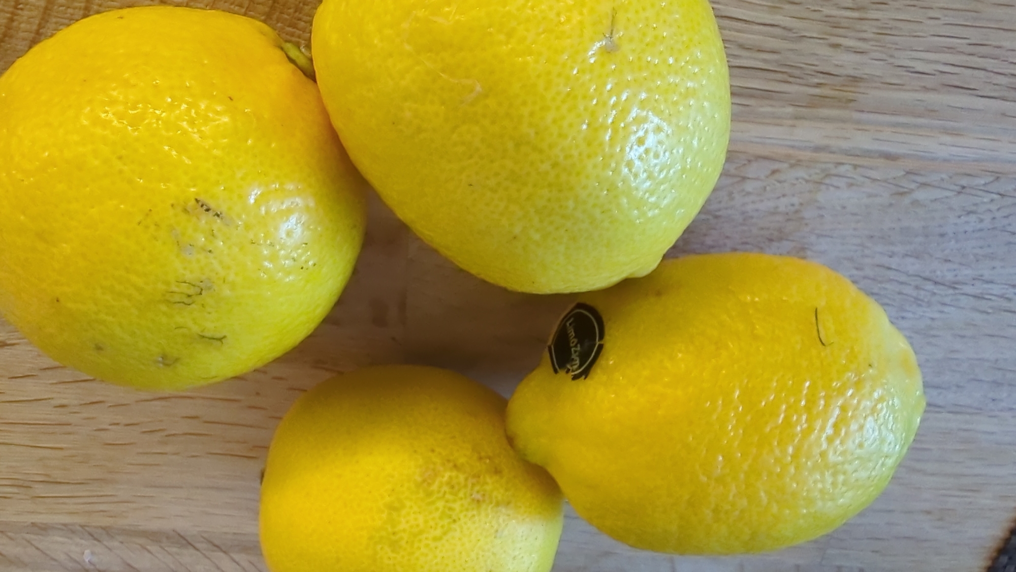 Four bright yellow lemons are arranged on a light-colored wooden surface. One lemon is in the upper left, another is in the upper right, and two are in the lower part of the image, one on the left and one on the right. The lemon in the upper left has a black sticker with white text, which appears to say "LimoZest". The surface has a visible wood grain pattern. The lighting suggests an indoor setting. There is no indication of the city of Kløfta, Norway, in the image.