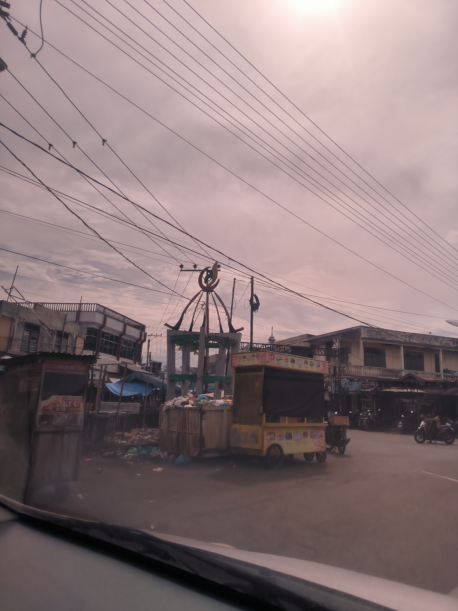 This image captures a street scene in Teungoh Baro, Indonesia, during an overcast day. The sky is a prominent feature, covered by a light pinkish-grey layer of clouds. Numerous dark power lines crisscross the upper portion of the frame, some with small insulators or bird-like objects attached.
In the midground, a bustling urban environment unfolds. Dominating the center is a decorative, possibly monument-like structure, featuring an ornate frame and topped with a large, green crescent moon and star symbol, suggestive of an Islamic context, perhaps a mosque finial or decorative archway. Around this structure and extending to the left, various makeshift stalls and market structures are visible. One prominent yellow food cart with wheels and a dark canopy sits on the right, displaying images of food products. To its left, another stall, partially obscured, shows text that includes "ZIBU KFC" and "KFC," indicating a fried chicken vendor. Piles of trash and debris are noticeable around these stalls and the central structure, hinting at a less regulated waste disposal system.
Several multi-story buildings, appearing somewhat aged with balconies and shuttered windows, line the street. One building on the left has white railings on its upper floor. On the right side of the street, a person is seen riding a motorcycle, suggesting daily transport and activity. The photograph itself is taken from inside a vehicle, with the top of a dashboard or car interior visible in the foreground at the bottom of the image. The overall scene depicts a functional, busy street with a blend of permanent structures and temporary market setups, under a muted, cloudy sky.