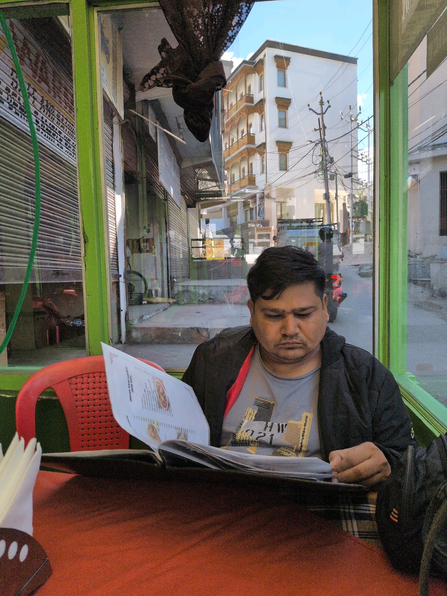 A man is sitting at a table in a cafe in Leh, India, looking at a menu. He is wearing a black jacket over a grey t-shirt with a graphic print. A red plastic chair is visible to his left, and a table covered with an orange cloth is in front of him. Through the cafe window, a street scene is visible, with buildings, power lines, and a motorcycle. The cafe has green window frames. The man's hands are on the menu, which is open. A black bag is partially visible on his right.