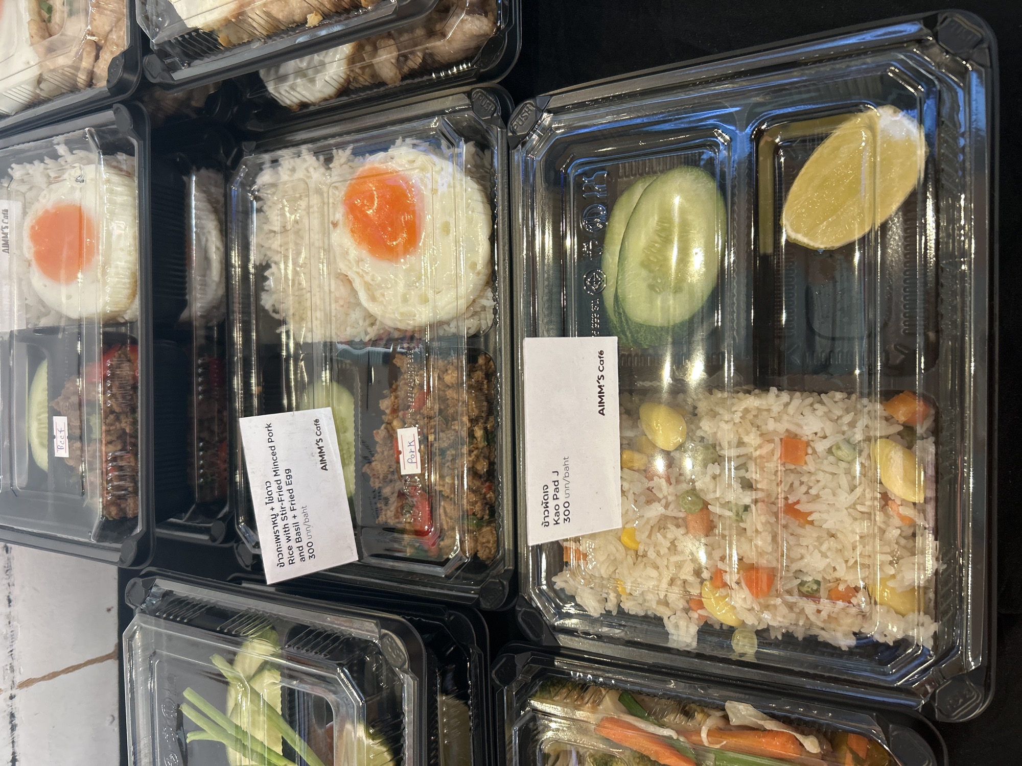 The image shows a close-up, overhead view of several take-out food containers arranged on a dark surface. The containers are clear plastic with black lids and appear to be stacked or placed next to each other.
In the foreground, a clear plastic container holds a portion of white rice mixed with various ingredients including diced carrots, green peas, corn kernels, and several large yellow ginkgo nuts. To the right of this container, a wedge of lemon is visible in a separate compartment of the same container.
Above and to the left of the rice dish, another clear plastic container is visible, containing a portion of stir-fried minced pork with basil and a fried egg. A white label attached to this container displays Thai script and reads, "ข้าวหมูสับ + ใบดาว Rice with Stir-fried Minced Pork and Basil + Fried Egg 300 บาท/baht AIMM's Cafe".
Further to the right, another take-out container shows a portion of rice topped with a fried egg. A small label on this container reads "Beef".
In the upper left corner of the image, a container with some greenery, possibly scallions, and a clear liquid is partially visible. To the right of this, another container holds a stir-fried meat dish, and a label reads "Pork".
The overall scene suggests a food vendor or restaurant setting in Tambon Mai Khao, Thailand, where these meals are prepared for take-out. The lighting is direct, highlighting the food items within the transparent containers.
