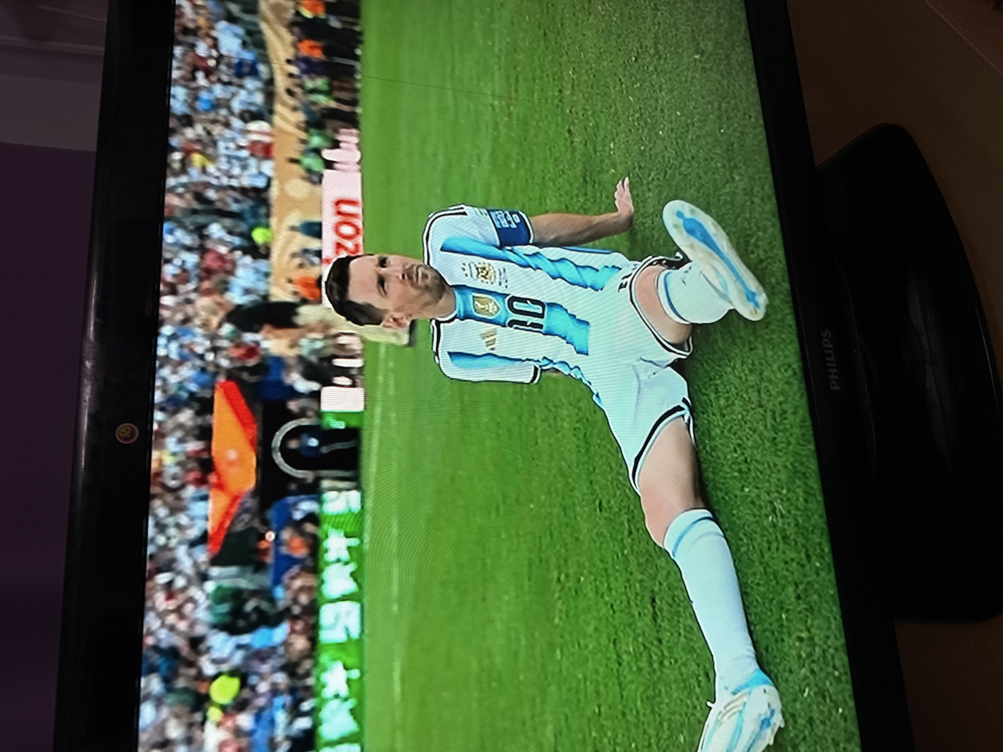 The image is a photograph of a television screen displaying a soccer match. On the screen, a male soccer player, identified as Lionel Messi by his jersey number '10' and facial features, is sitting on a vibrant green soccer field. He is wearing a white and light blue striped jersey and shorts, with white socks and light blue cleats. The player is looking to his left, with his right leg extended forward and his left leg bent. In the background, there are spectators in the stands, some holding flags, and advertising boards. The television screen is dark at the bottom with a visible "PHILIPS" logo. The scene is framed by the black bezel of the television set and a purple background above it. There is no indication of the city of Ufa, Russia in the image.