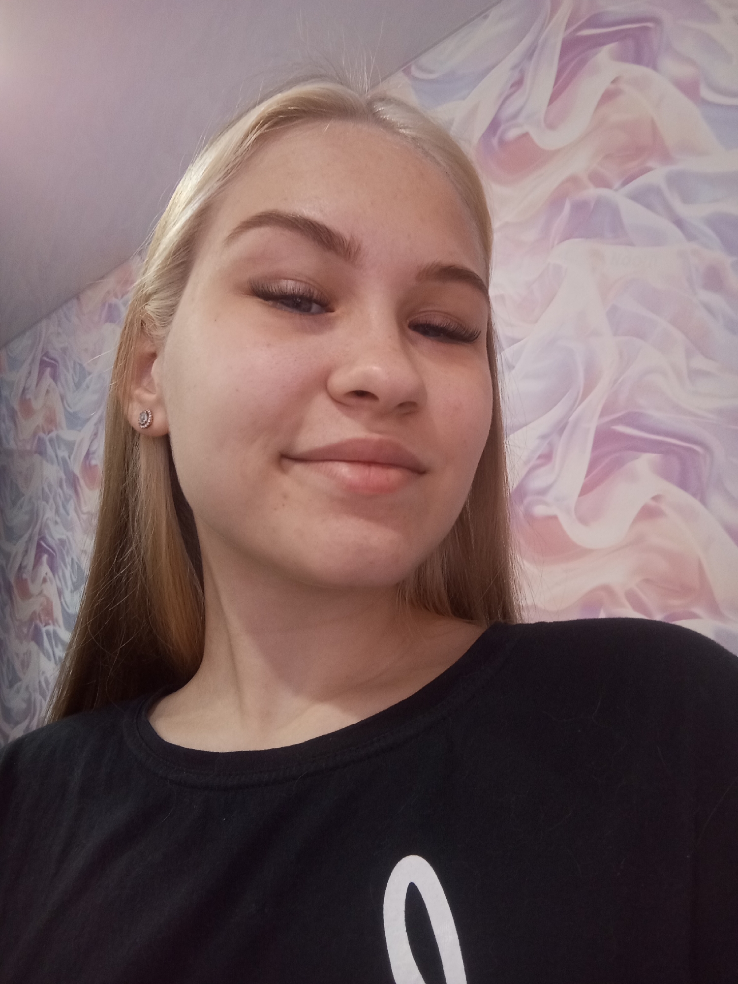 A young woman with fair skin, blonde hair, and long eyelashes is posing for a selfie indoors. She is wearing a dark t-shirt with a partial white design visible. Her expression is a slight smile, and her eyes are partially closed. A small stud earring is visible in her left ear. The background consists of a wall covered with a patterned wallpaper featuring a blend of pastel colors. It is not possible to determine the location in Krasnodar, Russia from the image alone.