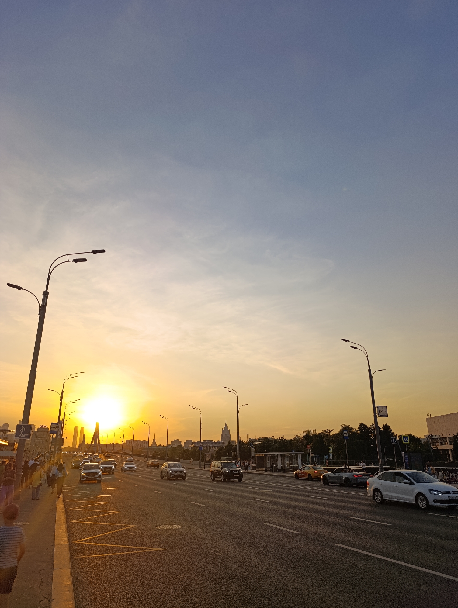 The image depicts a busy multi-lane highway or bridge in Moscow, Russia, at sunset, observed from a pedestrian sidewalk. The vantage point is likely looking west from the Krymsky Bridge (Crimean Bridge) over the Moskva River.
The sun, a brilliant yellow-orange orb, dominates the center of the midground, low on the horizon and creating significant glare and warm illumination across the scene. The sky transitions from a deep blue at the top to a vibrant orange and yellow near the horizon, with faint wisps of white clouds.
On the left, a concrete sidewalk accommodates approximately 10-15 pedestrians, most of whom are adults casually dressed, walking in various directions. In the immediate foreground, a young person with their back to the camera, wearing a blue and white striped shirt and dark shorts, walks away from the viewer. Tall, dark grey street lamps with multiple light fixtures line both the sidewalk and the road. A blue pedestrian sign with a white walking figure is visible on the left. Further down, an "AKKO" sign is affixed to a building or structure.
The multi-lane asphalt road occupies the right and central portions of the image, showing moderately heavy traffic. Numerous vehicles, predominantly cars (sedans, SUVs, a convertible, and a yellow taxi) in shades of white, silver, and dark grey, travel away from the camera. Notable vehicles include a white Volkswagen Polo in the rightmost lane and a grey convertible in the middle lane. The road features white dashed lane dividers and distinctive yellow zigzag markings on the shoulder, potentially indicating a bus stop or no-parking zone. A street lamp on the right side of the road features a rectangular clock.
In the distant background, the Moscow cityscape is visible. To the left of the sun's intense glare, modern glass skyscrapers, characteristic of the Moscow International Business Center ("Moscow City"), pierce the sky. To the right, a distinctive Stalinist-era skyscraper with a prominent spire, identifiable as the main building of Moscow State University (MGU) on Sparrow Hills, rises above the horizon. Patches of green foliage from trees are visible along the sides of the road.