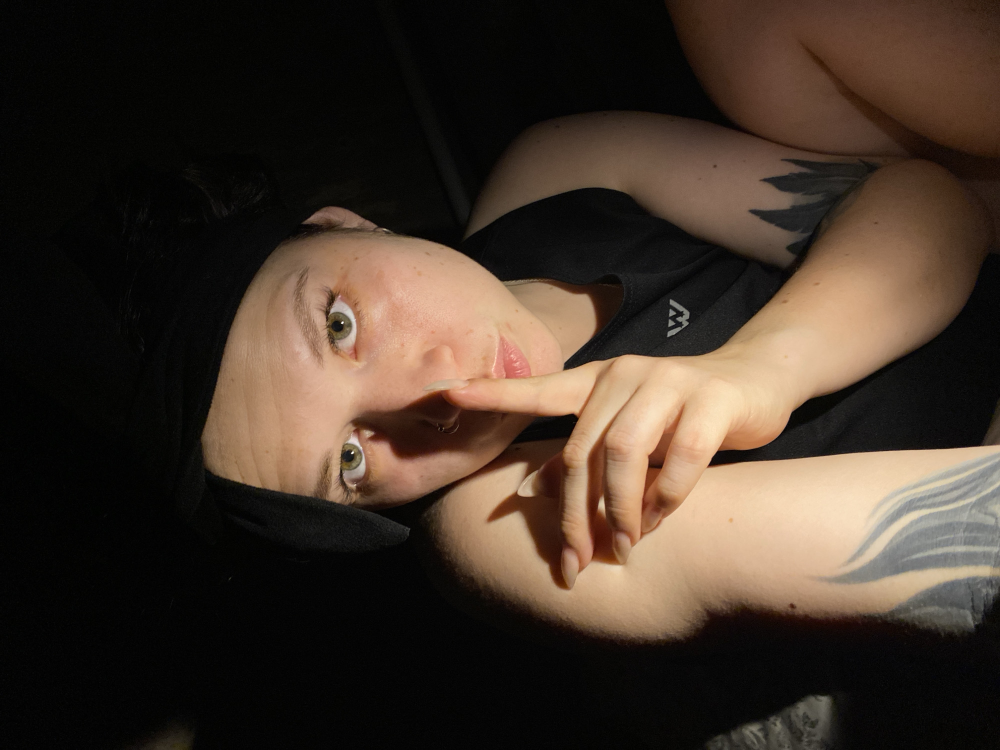 A person with a black headband is lying down in dim lighting, holding a finger to their lips in a "shhh" gesture. They are wearing a black tank top with a white logo. Tattoos are visible on their arms. The setting appears to be indoors. There is no indication of the location being in Chaikovsky, Russia.