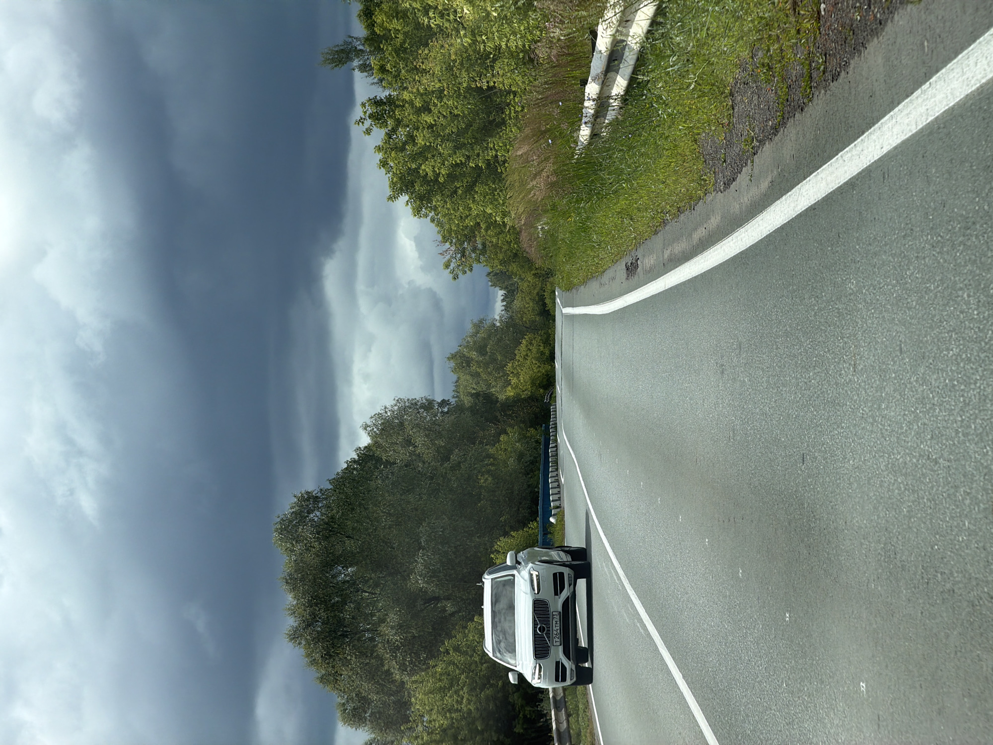 A white Volvo XC90 SUV is driving on a paved road with a white dividing line. The road curves to the right, bordered by a metal guardrail and lush green vegetation on the right side of the frame. On the left side, a dense line of trees and foliage stretches into the distance. The sky above is filled with dramatic, dark grey clouds, suggesting an impending storm. The scene appears to be in a rural or semi-rural area, possibly on the outskirts of Staryi Urenbash, Russia, given the provided location context. There are no people visible in the image. The SUV's headlights are illuminated.