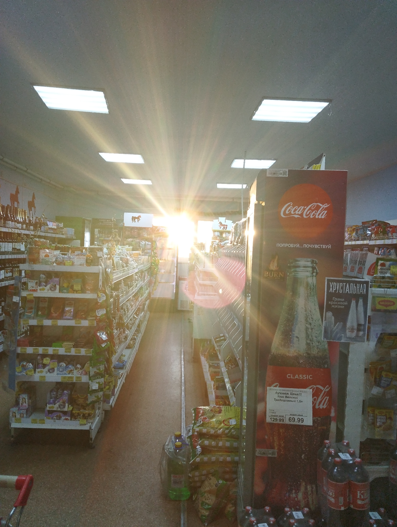 The image shows an aisle in a store, likely a grocery store, in Krasnogorskoye, Russia. Shelves filled with various products line both sides of the aisle. A Coca-Cola advertisement, featuring a bottle of classic Coca-Cola and Russian text, is prominently displayed in the foreground, along with a price tag of 69.99. There are packs of water and bags of crisps by the Coca-Cola advertisement. Sunlight streams through the far end of the aisle creating a bright glare. Fluorescent lights are visible in the ceiling. A red shopping cart is partially visible in the bottom left corner.