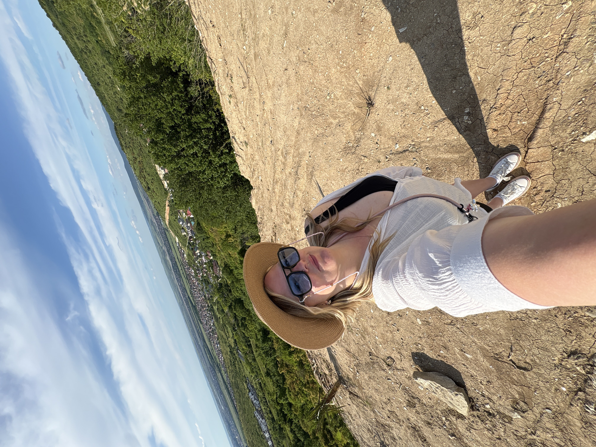 This image, rotated 90 degrees counter-clockwise, depicts a woman taking a selfie from an elevated viewpoint on a bright, sunny day. The woman has blonde hair and is wearing a white textured long-sleeved top, a straw hat, and dark sunglasses with a light-colored chain. A black bra strap is visible beneath her top, and a pink strap, possibly from a bag, crosses her chest. Her white sneakers with reddish-brown soles and white socks are visible as she stands on dry, cracked, light brown earth. A long, distinct shadow of her legs and body is cast on the ground, suggesting either late morning or late afternoon sunlight.
The setting is an expansive outdoor landscape. To the woman's left, a dense, green forest covers a steep slope that descends into a valley. In the far distance, a small town or village with numerous buildings is visible amidst the verdant scenery, stretching towards the horizon. The sky above is a clear blue with scattered white, wispy clouds. The scene conveys a sense of a peaceful, warm day with a breathtaking panoramic view. No discernible text is visible in the image.
