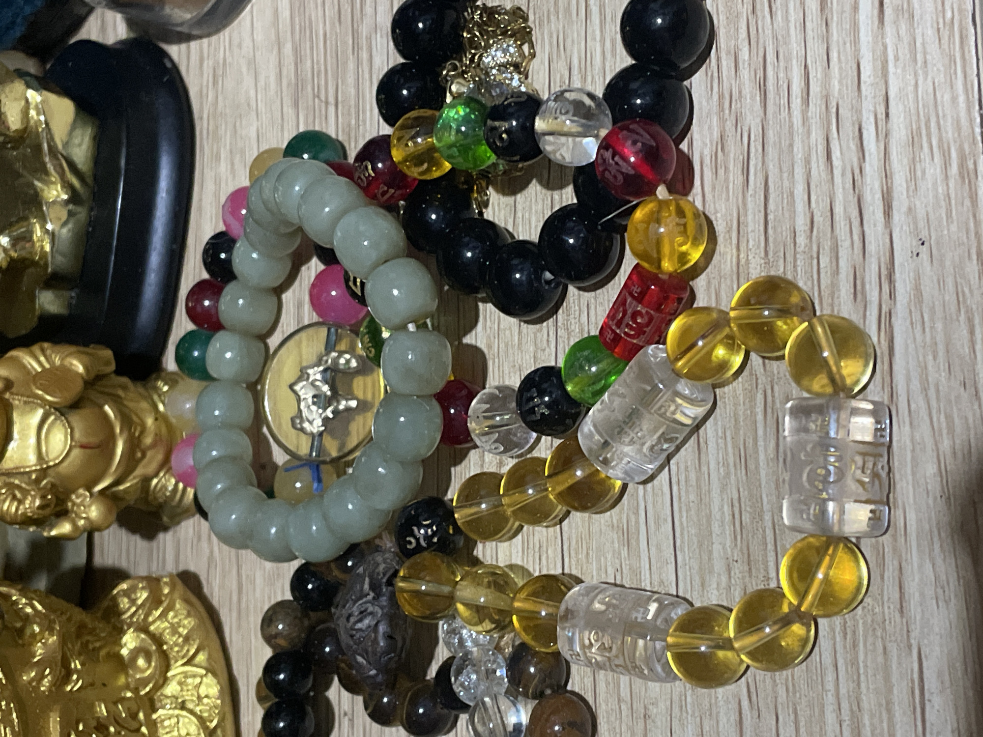 The image displays a collection of beaded bracelets and two small golden figurines on a wooden surface. One figurine is a seated Buddha-like figure with a serene expression, and the other is a rounder, jovial-looking figure, possibly a deity. The bracelets are varied in color and material, including light green jade-like beads, yellow translucent beads, black beads, clear beads with inscribed Chinese characters, and red and pink beads. Some bracelets feature charms, such as a coin-shaped charm with an animal engraving. The overall scene is a close-up shot of these decorative items, with the focus on the textures and colors of the beads and figurines. The image does not contain any specific landmarks or indicators that would allow for the precise identification of its location within Davao City, Philippines.