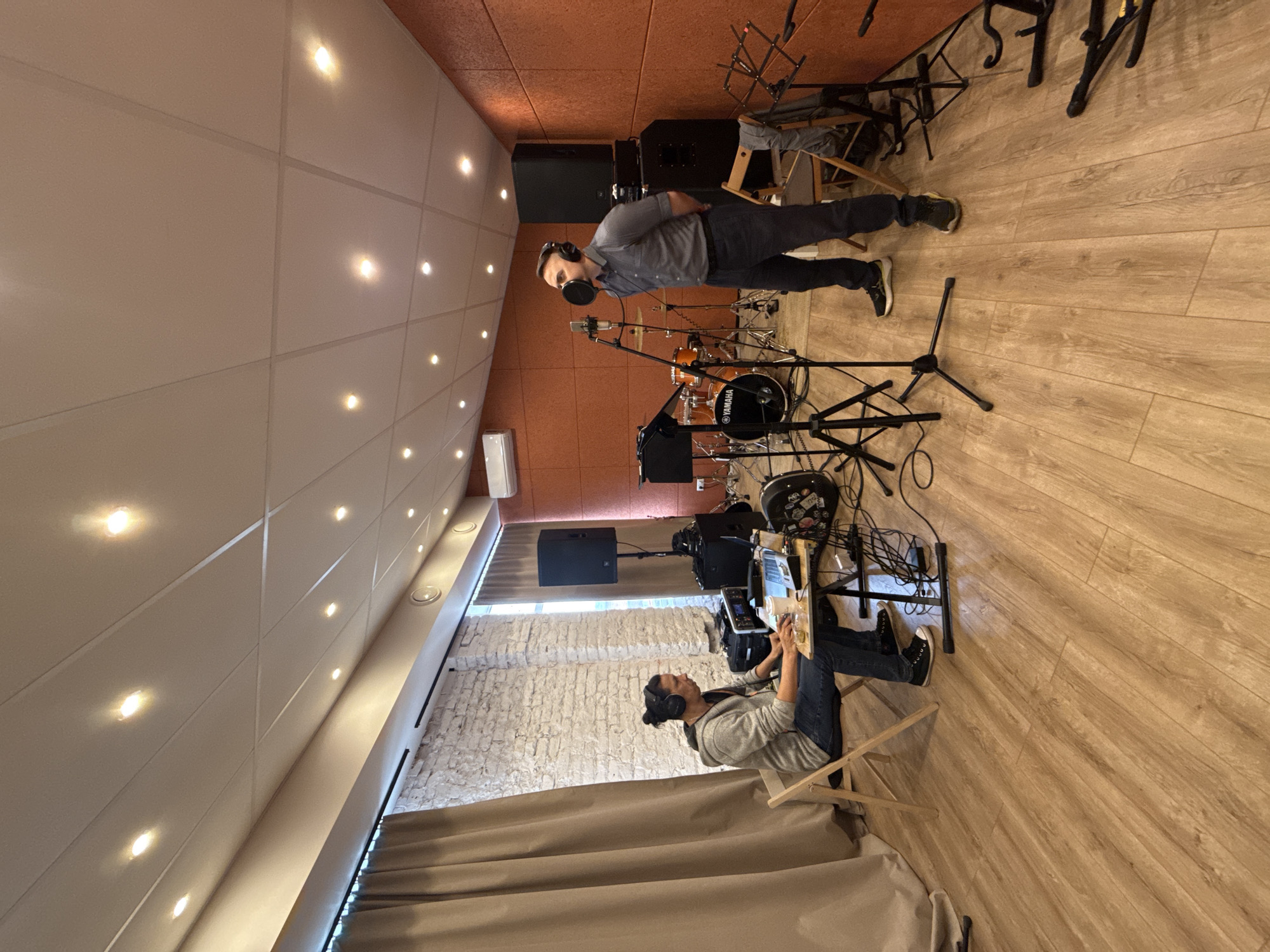 The image shows the interior of a recording studio. Two men are present. One man is standing in front of a microphone, wearing headphones, and possibly singing or speaking. He is dressed in a gray shirt and dark pants. The second man is sitting on a folding chair, also wearing headphones, and appears to be working on a laptop or similar device. The studio contains a drum set and several pieces of audio equipment, including speakers and microphone stands. The walls are a mix of brick and acoustic paneling. The ceiling has recessed lights. The location is assumed to be a recording studio in or near Bobrovo, Russia, although there's no explicit geographical information in the image.