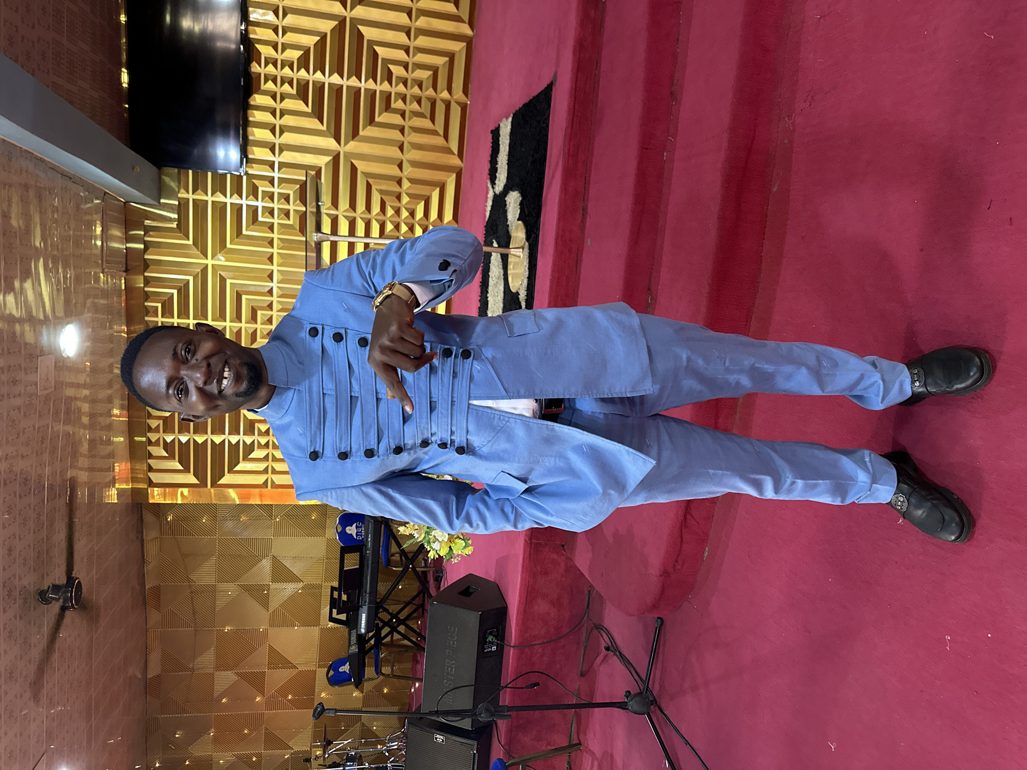 A man stands on a red carpeted stage in what appears to be a church or auditorium in Narayi, Nigeria. He is wearing a light blue suit with a high collar and decorative straps across the chest. The background features a textured golden wall with a geometric pattern, and a black and white rug is visible on the stage. To the left, musical instruments including a keyboard and speakers are set up. The man is smiling and pointing with his right index finger towards the viewer.