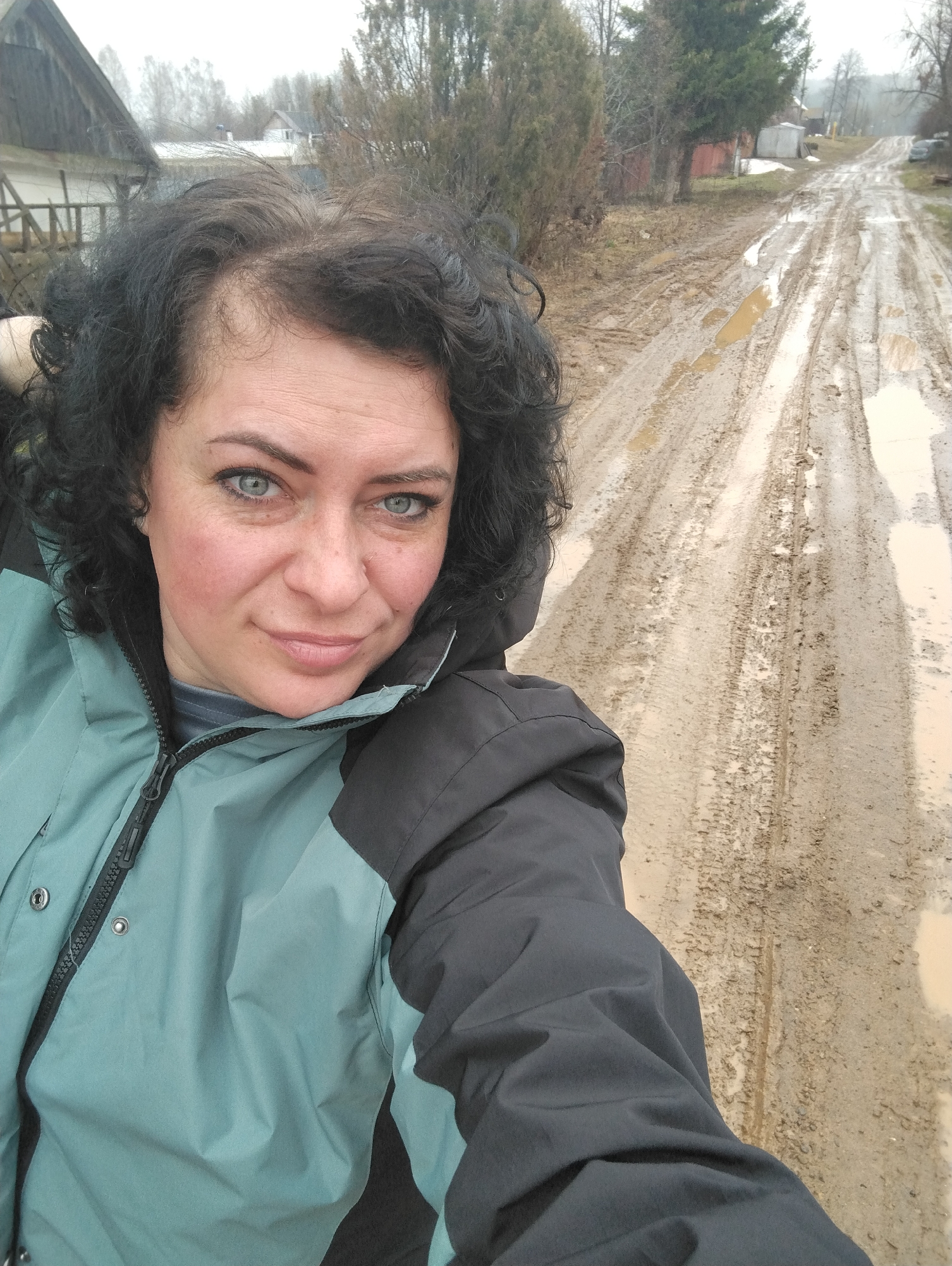 The media file features a close-up selfie of a woman outdoors in a rural setting, specifically Grishanki, Russia. The woman, with dark, curly hair and light blue eyes, occupies the left side of the frame, looking directly at the camera with a neutral to slightly pleasant expression. Some gray hairs are visible on her scalp near her forehead. She is wearing a practical two-tone jacket, primarily teal or bluish-green on the main body and dark grey or black on the sleeves and shoulders, with a visible zipper down the front.
Behind and to the right of the woman is a deeply rutted and extremely muddy dirt road, indicating recent heavy rain or a thaw. Large puddles of murky water are scattered along the tire tracks. The road stretches into the distance, flanked by sparse trees and vegetation, mostly bare of leaves, suggesting a cold or transitional season. In the background, beyond the trees, several simple, somewhat rustic houses or buildings with dark roofs are visible under an overcast sky. The overall atmosphere suggests a damp, perhaps chilly, daytime. There is no visible text in the image.