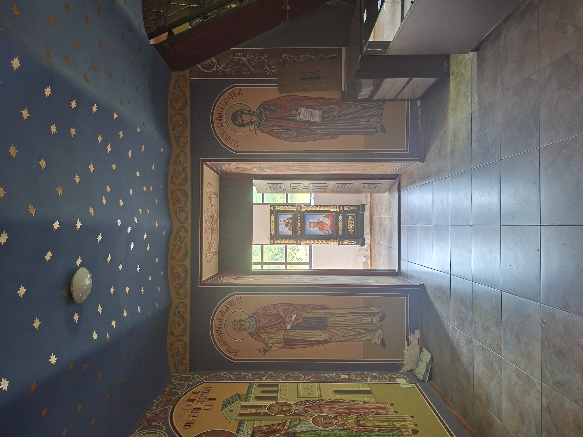 The image shows the interior of an Orthodox church in Condrița, Moldova. The ceiling is painted dark blue and adorned with small, gold star-like decorations. A central light fixture is visible on the ceiling. The walls are decorated with religious icons painted in a traditional style.
On the left wall, a large icon depicts several figures, including one labeled "Sf. Voievod Constantin Brancoveanu cu fiii". In the center of the image, an arched doorway leads to another part of the church. On either side of this doorway, within arched niches, are large icons of saints. The icon on the left depicts a bearded male saint holding a scroll. The icon on the right depicts a female saint with a veil, holding a cross and a scroll.
In the doorway, a triptych icon is visible in the distance, depicting religious figures. The floor is tiled with large, square, light-colored tiles. To the right of the central doorway, a dark wooden structure with metal accents is partially visible, possibly part of a display or altar.
The overall impression is of an ornate, sacred space with rich religious artwork. The lighting suggests it might be daytime, with some natural light entering from the doorway.