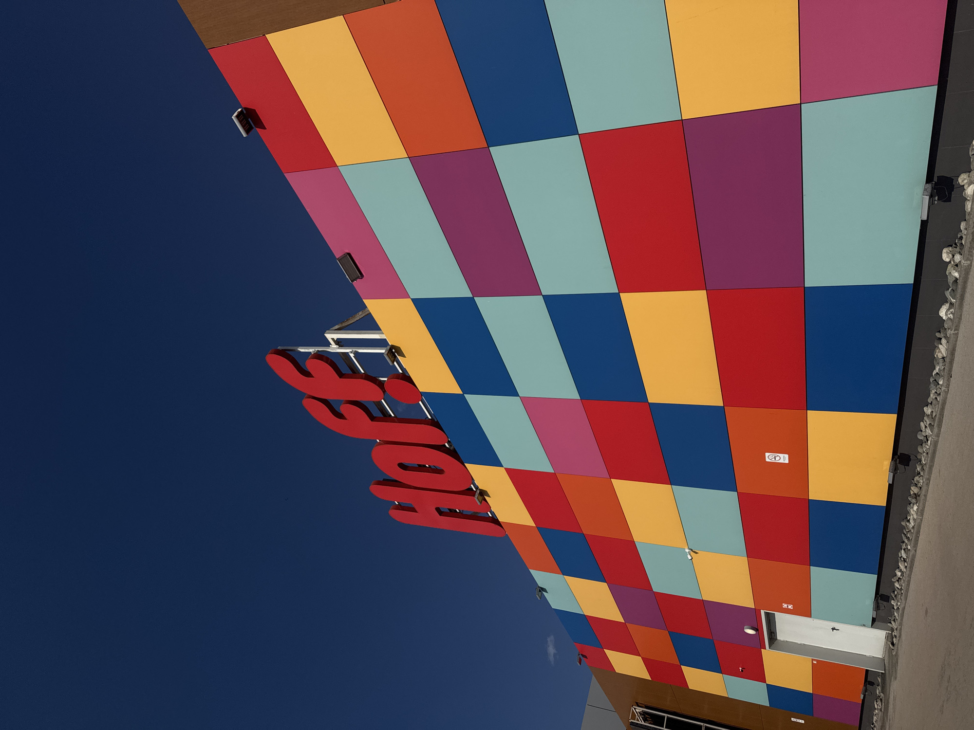 The image depicts the exterior of a building in Unknown, Unknown. The building has a facade composed of colorful square panels arranged in a checkered pattern. Colors include red, orange, yellow, light blue, dark blue, and purple. The name "Hof" in large, red letters is attached to the building. The sky is a deep blue, visible in the upper left portion of the image. A white door is located at the bottom of the facade. The camera angle is from below, looking up at the building.