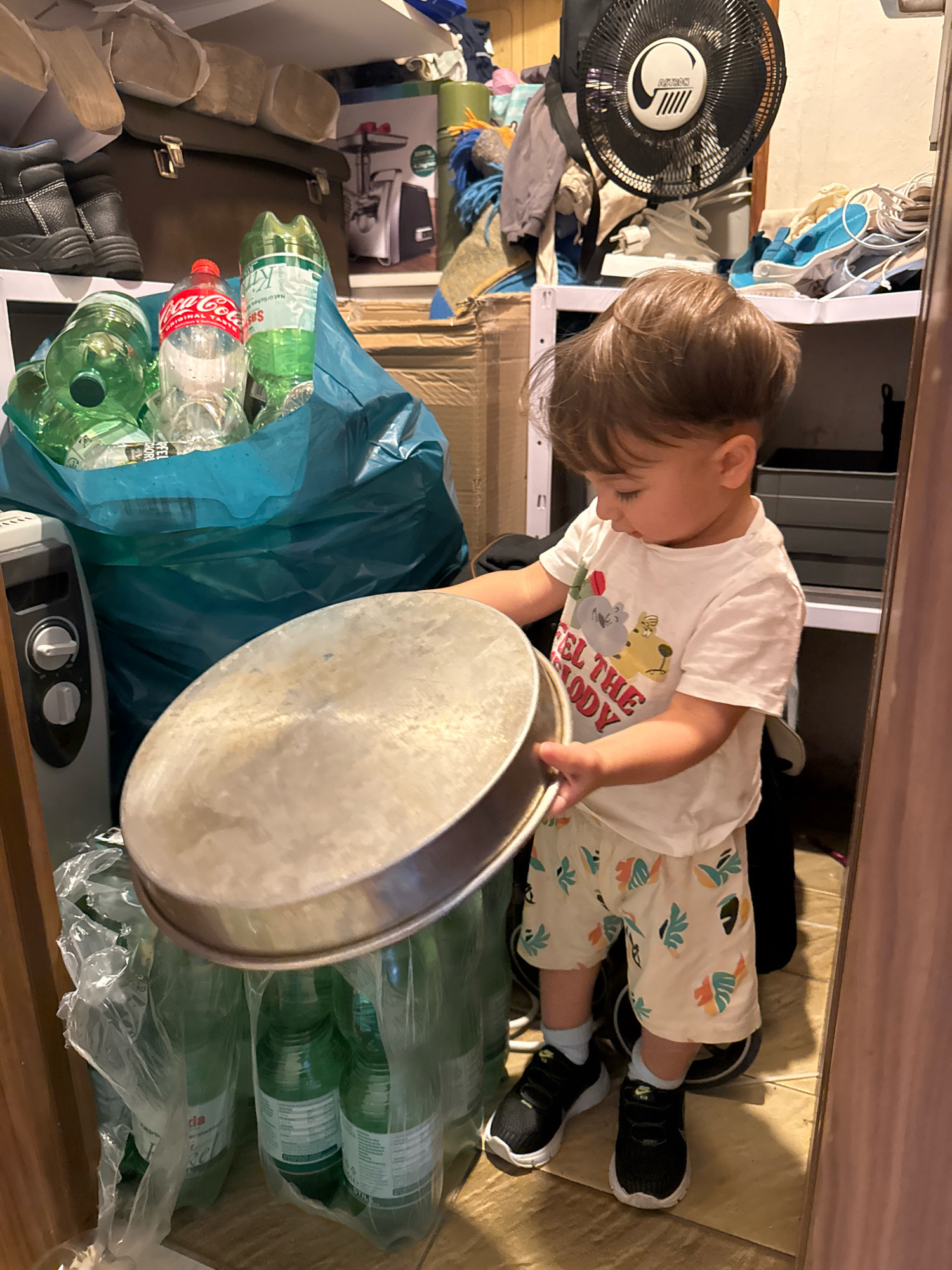 The image depicts a young child, likely a toddler, standing in a cluttered storage area. The child is wearing a white t-shirt with a graphic print and patterned shorts, along with black sneakers. The child is holding a large, circular metal tray and appears to be interacting with a large blue bag filled with plastic bottles.
The storage area appears to be a residential space, possibly a basement or a utility room, with shelves filled with various items, including boxes, clothing, shoes, and a fan. A radiator is visible on the left side of the frame. The environment is dimly lit and appears disorganized, suggesting it's a functional space rather than a display area. The overall scene captures a moment of a child's exploration within this environment.