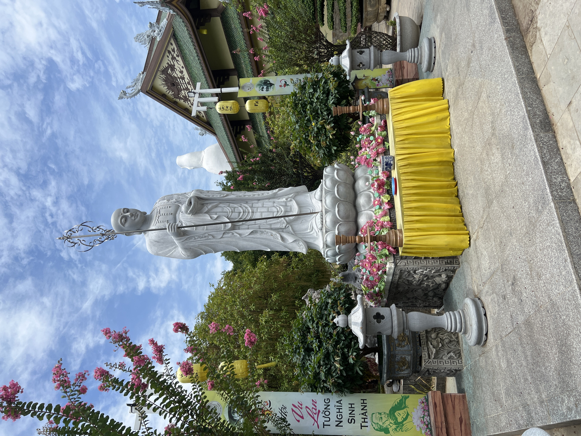 A large, white, stone statue of a Buddhist figure stands prominently in the foreground, facing forward and holding a staff. The statue is situated on a lotus flower base, which rests on a raised platform adorned with pink flowers and a bright yellow tablecloth. To the left and right of the statue are stone lanterns. In the background, a traditional Vietnamese temple with a green, tiled roof and ornate decorations can be seen. The sky above is partly cloudy with streaks of white clouds against a blue backdrop. The scene appears to be an outdoor courtyard within a temple complex, likely located in the city of Đà Nẵng, Vietnam, given the presence of Vietnamese text on a banner to the left of the statue. There are no people visible in the image. The overall atmosphere is serene and spiritual.