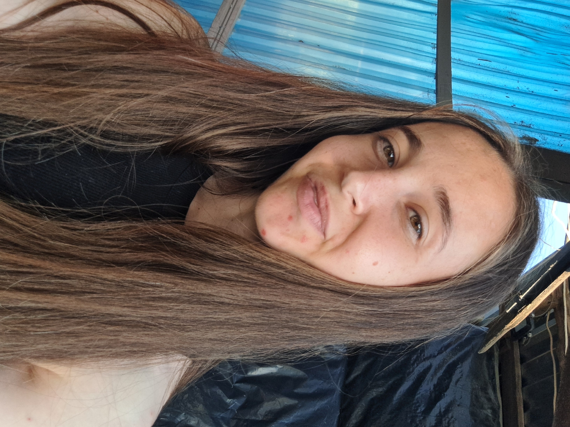 A young woman with long, brown hair is posing for a selfie in an outdoor setting. She is looking at the camera with a slight smile. The background is a textured blue corrugated surface, with some wooden beams and a black tarp visible. The scene appears to be located in the village of Vypasne, Ukraine.