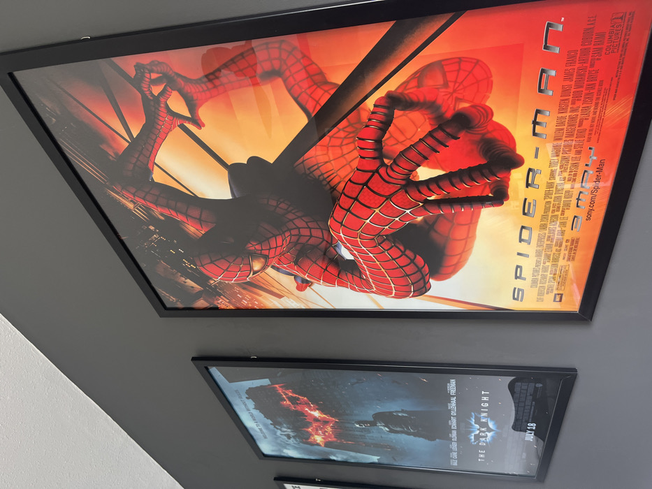 A framed "Spider-Man 2" movie poster features Spider-Man prominently in the foreground against an orange backdrop with a cityscape. Text along the right side includes cast and crew information, the title "Spider-Man 2", and the Sony Pictures logo. Below, a "The Dark Knight" poster is visible with cast members' names. Both posters are framed in black and hung on a grey wall. The view is a low-angle shot, slightly rotated.