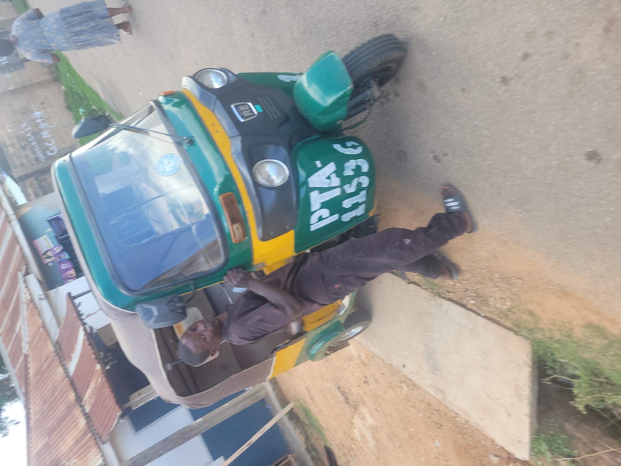 In Jos, Nigeria, a man in a dark t-shirt and black pants is seen leaning on a green and yellow auto-rickshaw. The auto-rickshaw has "PTA-11536" written in white on its front. The man appears to be a driver or mechanic. In the background, a woman in a blue patterned dress is walking away from the camera. Buildings with corrugated metal roofs are visible, along with some graffiti on a wall that reads "AVURIGADA 27 GCM 444". The ground is a mix of dirt and paved surfaces, with a concrete drainage channel on the left. The scene suggests a typical street environment in a Nigerian city.