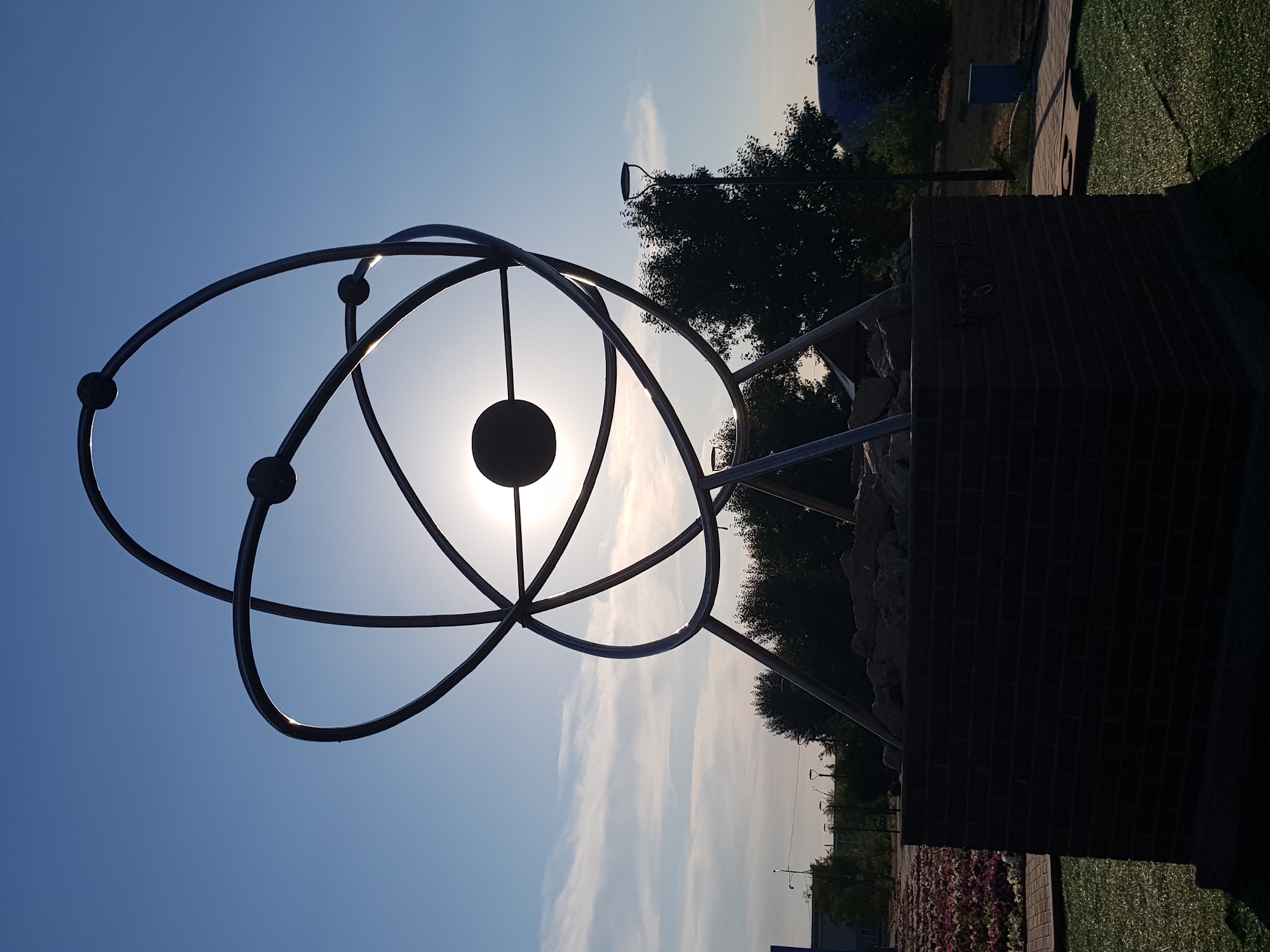 The image depicts an outdoor sculpture resembling an atomic model, with interconnected metallic rings and spheres, set against a bright sky. The sculpture is mounted on a brick pedestal, which is filled with rocks at the top. In the background, there are trees, streetlights, a pathway, and a flowerbed. The scene appears to be in a public space, possibly a park or plaza. The location is identified as the city of Kurchatov, Kazakhstan. The lighting suggests it is either early morning or late afternoon, with the sun partially obscured by the sculpture.