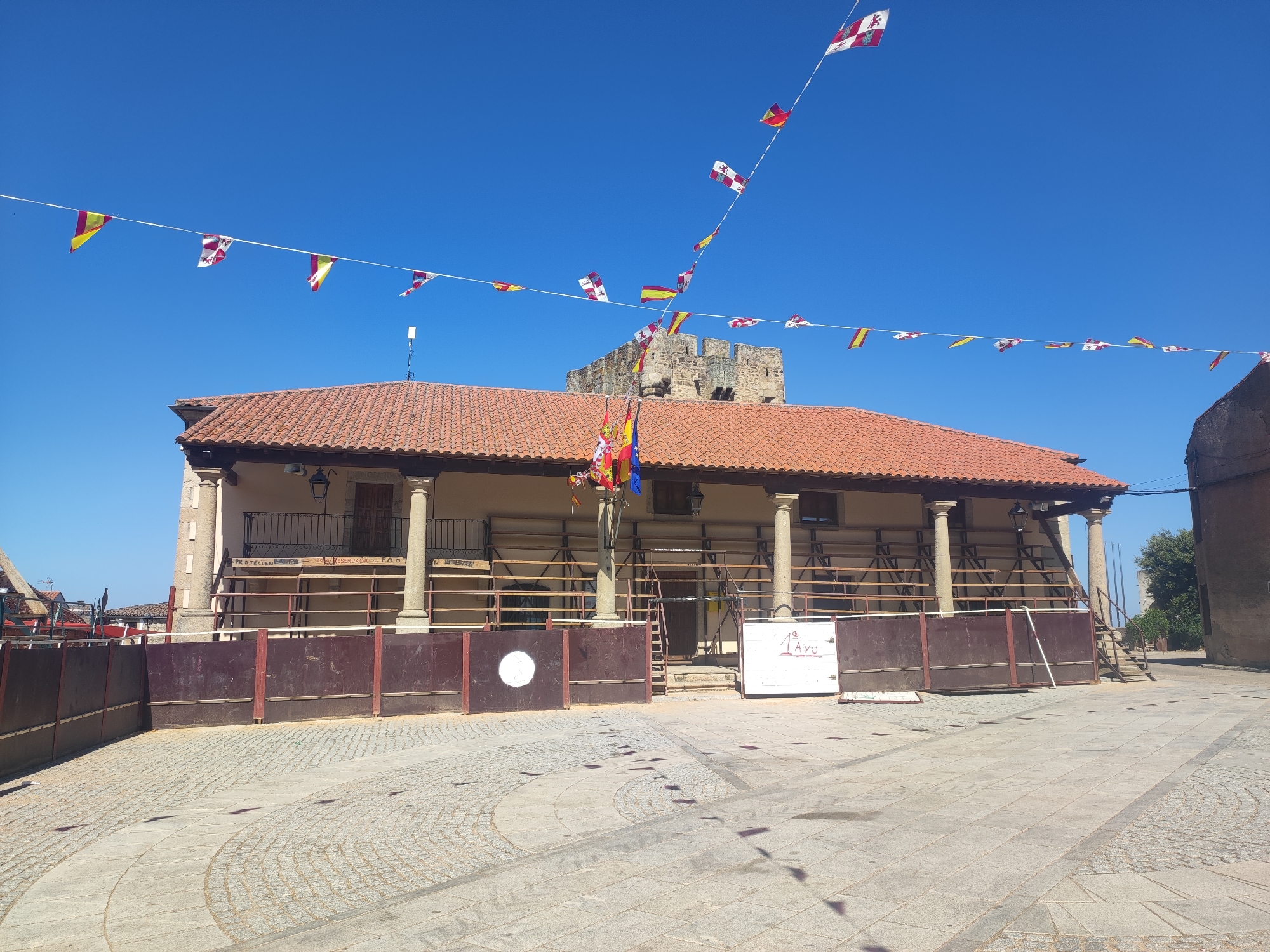 The image depicts an outdoor scene in Sobradillo, Spain, under a clear blue sky. In the foreground, a cobblestone plaza is visible, with some circular patterns etched into the paving. A dark brown, low fence or barricade runs across the middle ground. Behind this, a building with a red-tiled roof and a covered walkway supported by columns is present. This structure appears to be a temporary arena or bullring, as suggested by the fencing and scaffolding. Various flags, including the Spanish flag and other regional banners, are strung across the sky and on poles, indicating a festive occasion. In the background, a stone structure, possibly a tower or castle ruins, can be seen. The scene is devoid of people.
