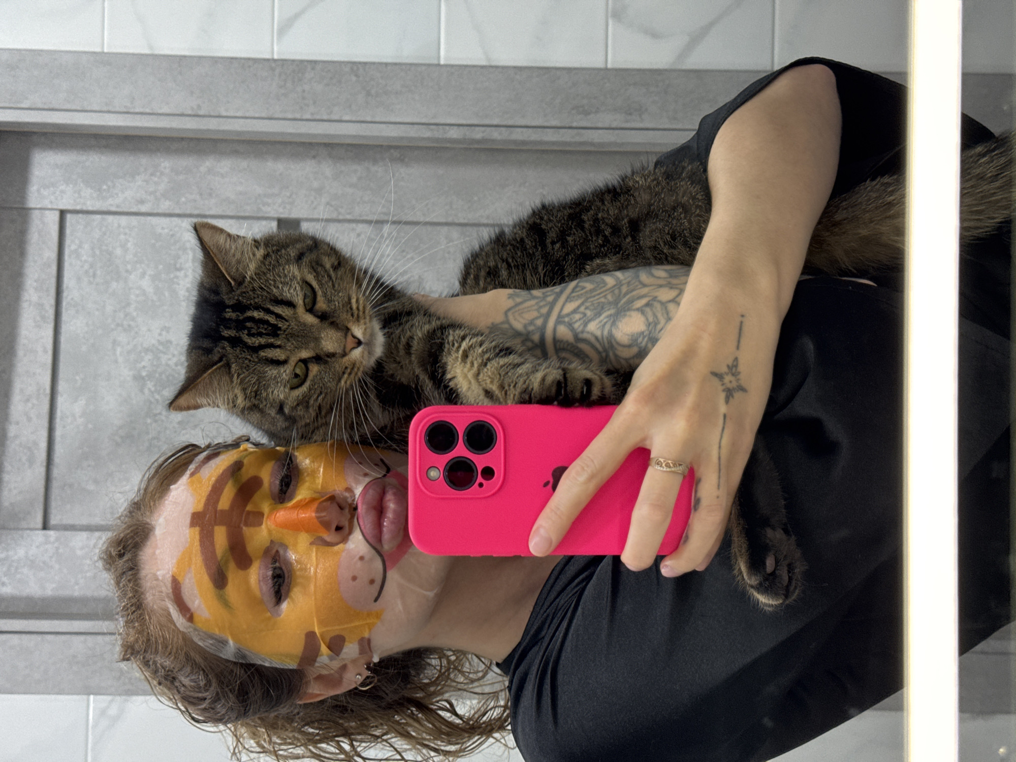 A person wearing a tiger-themed face mask is holding a pink iPhone to take a selfie. A brown tabby cat is draped over their left arm. The person's left arm is tattooed with geometric designs. Their ring finger has a gold ring. The background appears to be a bathroom with gray tiled walls. A partially illuminated light fixture is visible on the right side of the frame. The location is Chaikovsky, Russia.